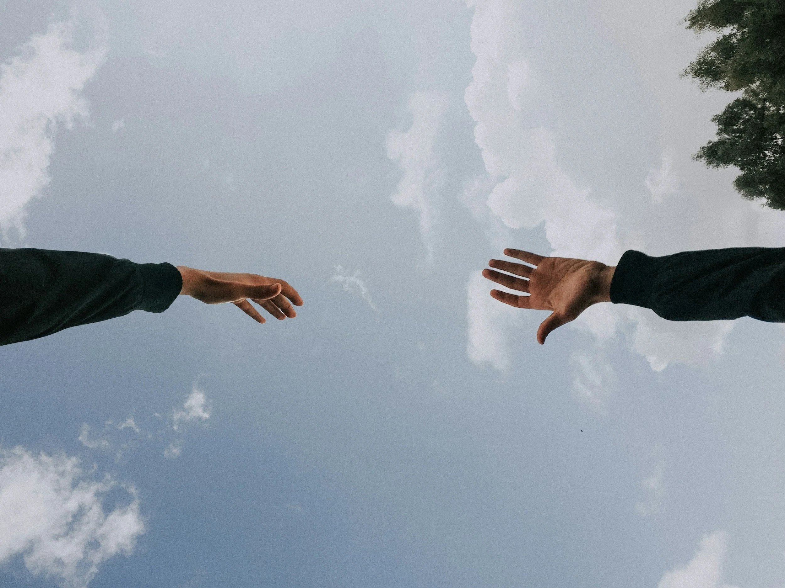 Two people reaching out their hands towards each other against a cloudy sky with trees on the sides.