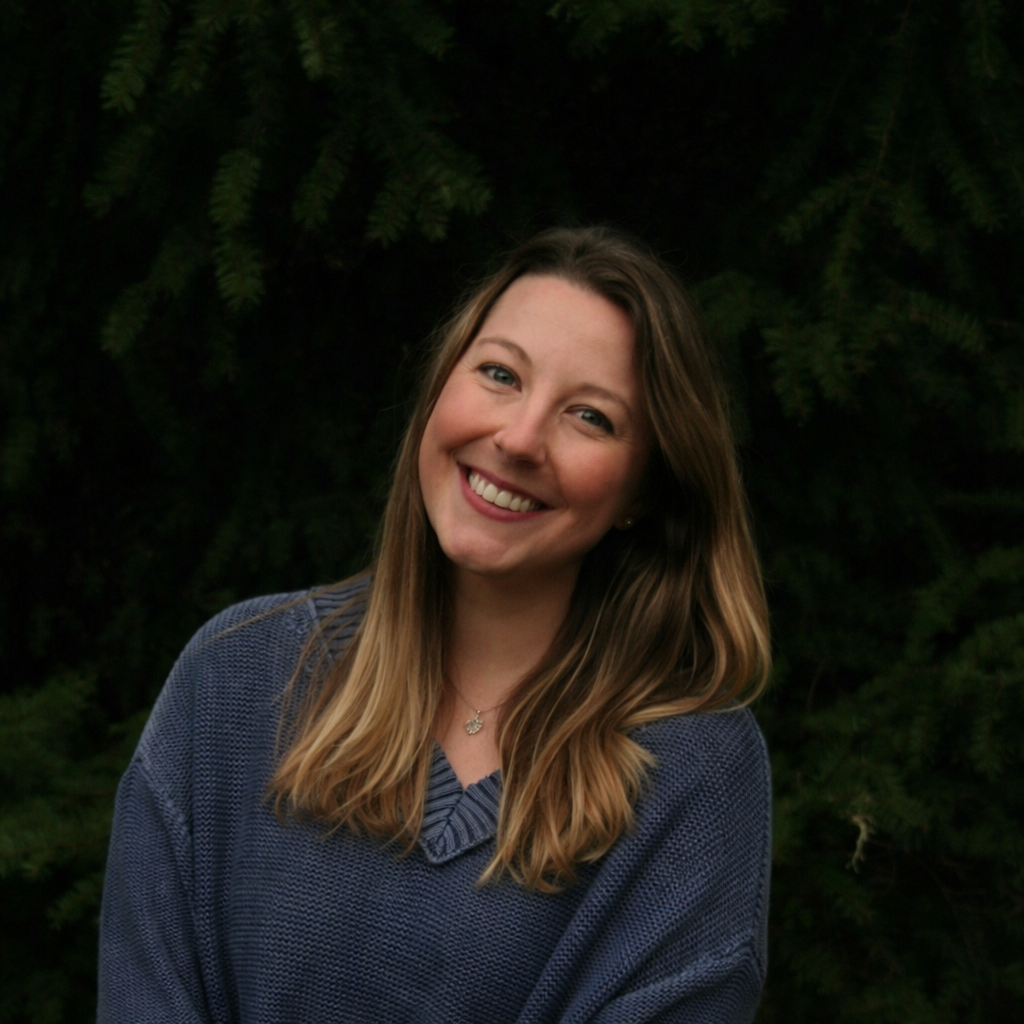 Smiling woman with long, wavy, light brown hair wearing a navy blue sweater, outdoors against a background of green foliage.