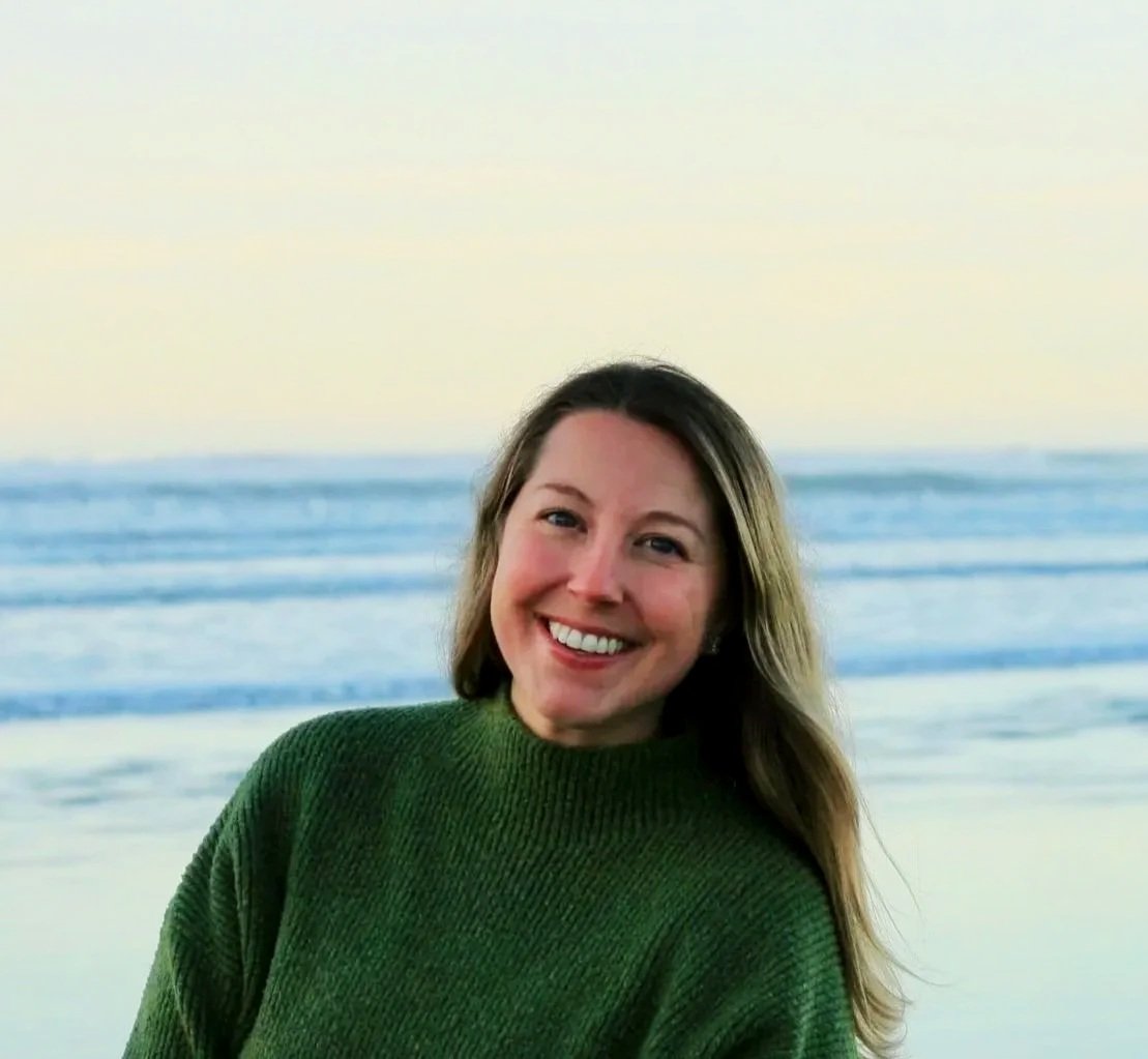 Sydney with long hair smiling at the camera on a beach at sunset, wearing a green sweater.