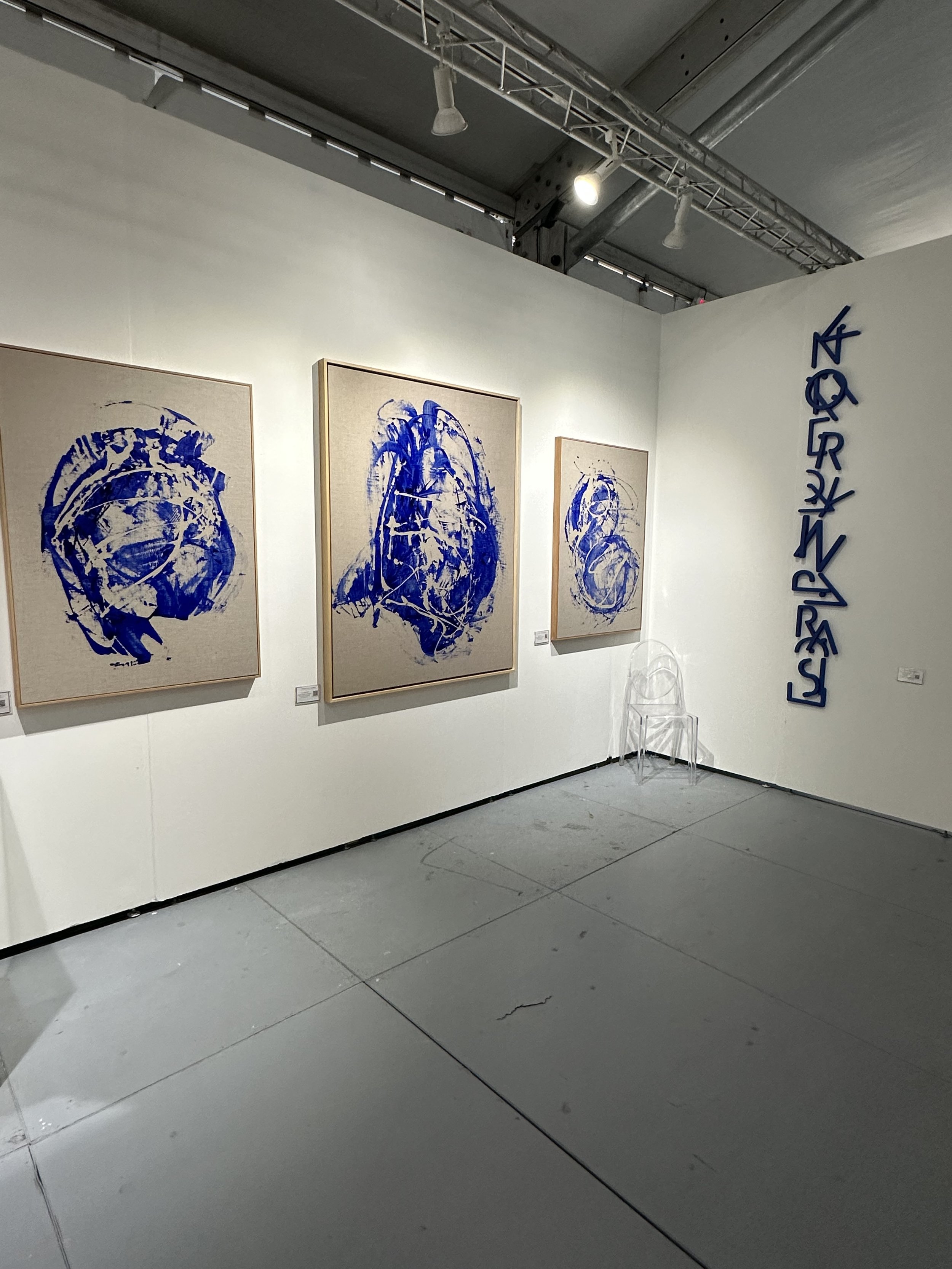 Abstract art gallery with three blue and white paintings on the wall, alongside blue abstract sculptures and a clear chair.