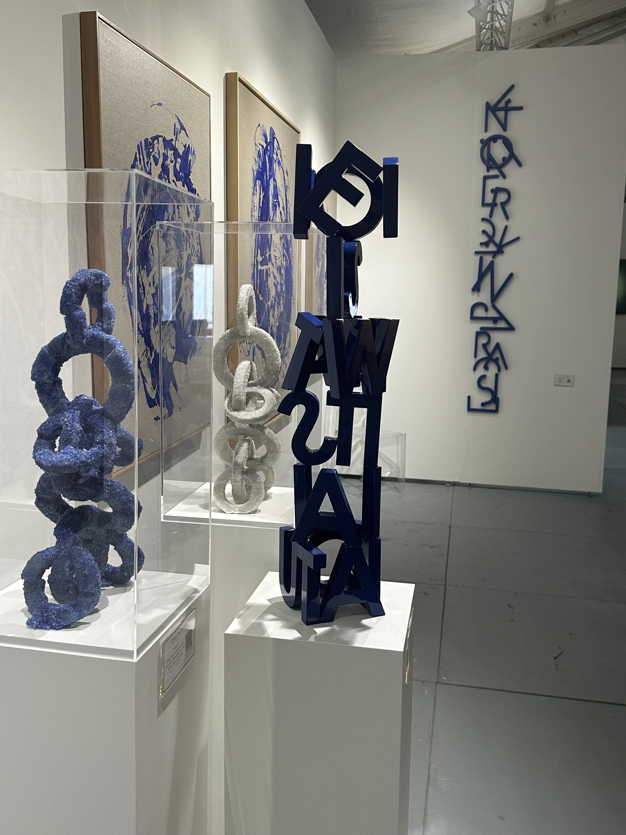 Contemporary art exhibit featuring abstract sculptures and wall art with geometric and letter-like elements, primarily in blue tones.