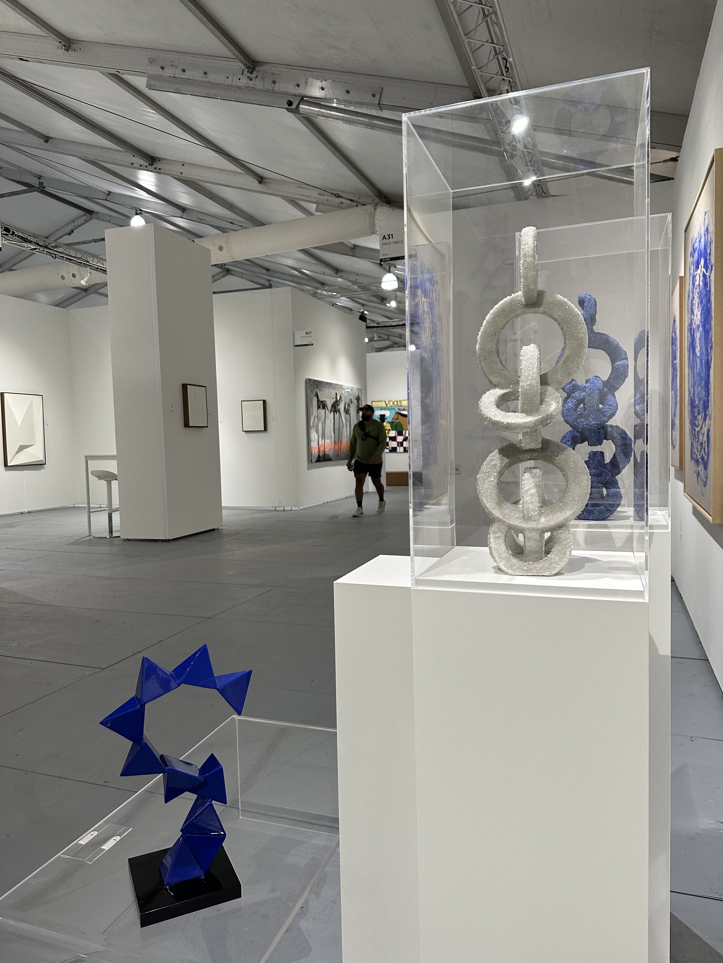 Contemporary art gallery interior with sculptures and paintings displayed. A foreground sculpture features geometric shapes in blue, while another piece in a glass case displays interconnected rings. Walls showcase various framed artworks.