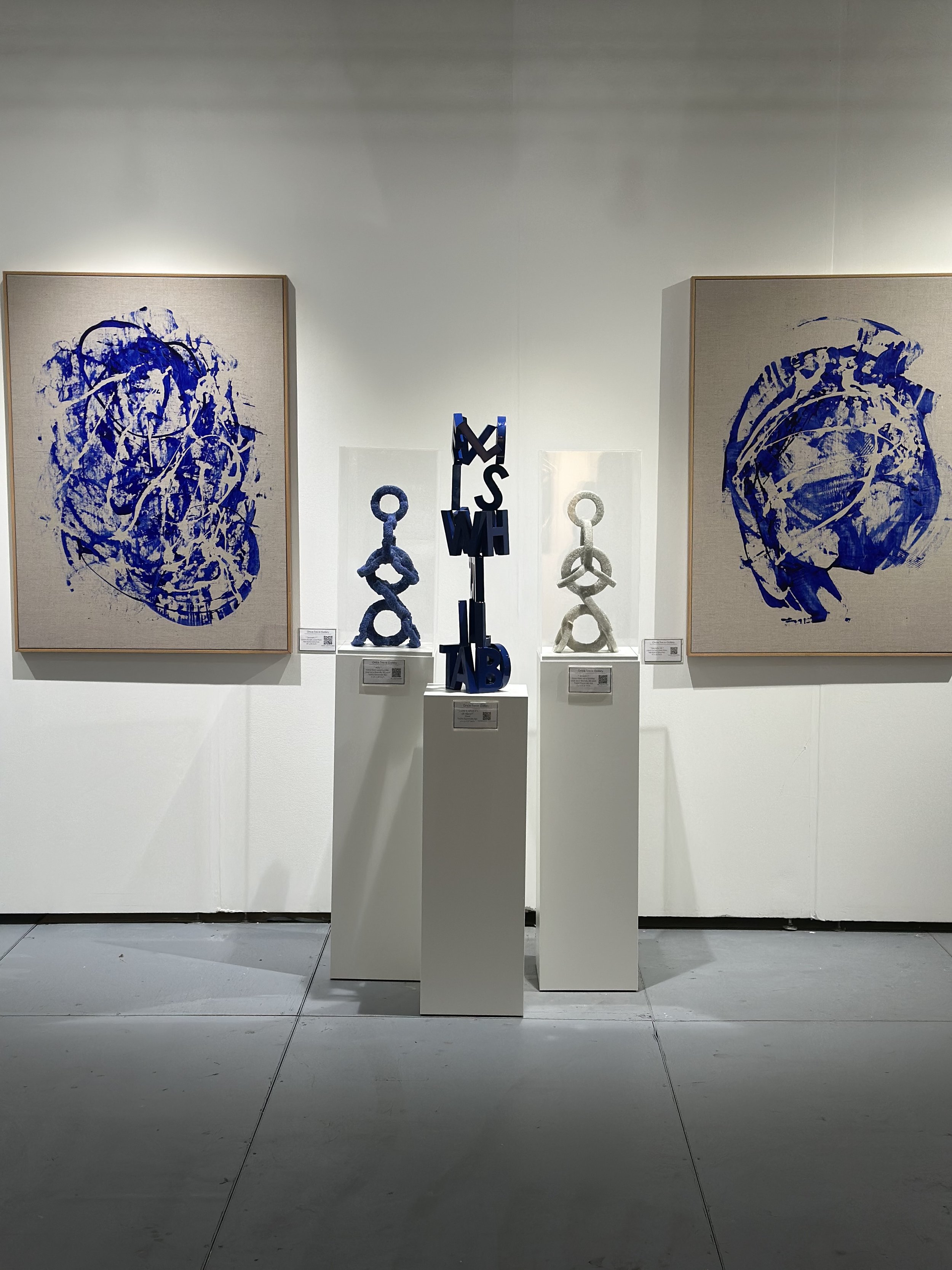 Art installation featuring two abstract blue paintings on canvases and three sculptural pieces, including one with stacked letters, displayed on white pedestals.