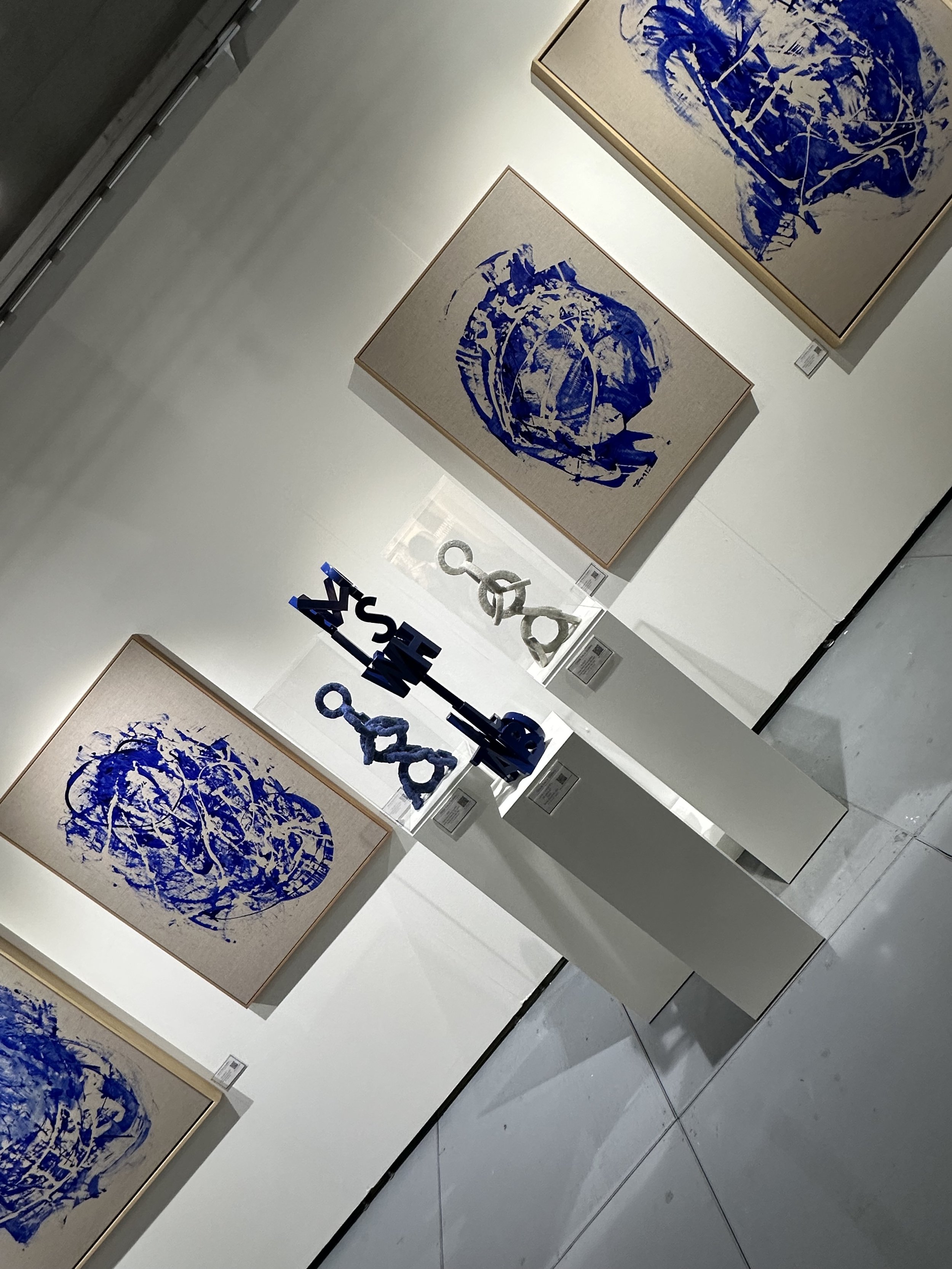 Art gallery exhibit showing three abstract paintings with blue splatters on canvas and three sculptures on pedestals, featuring blue elements.