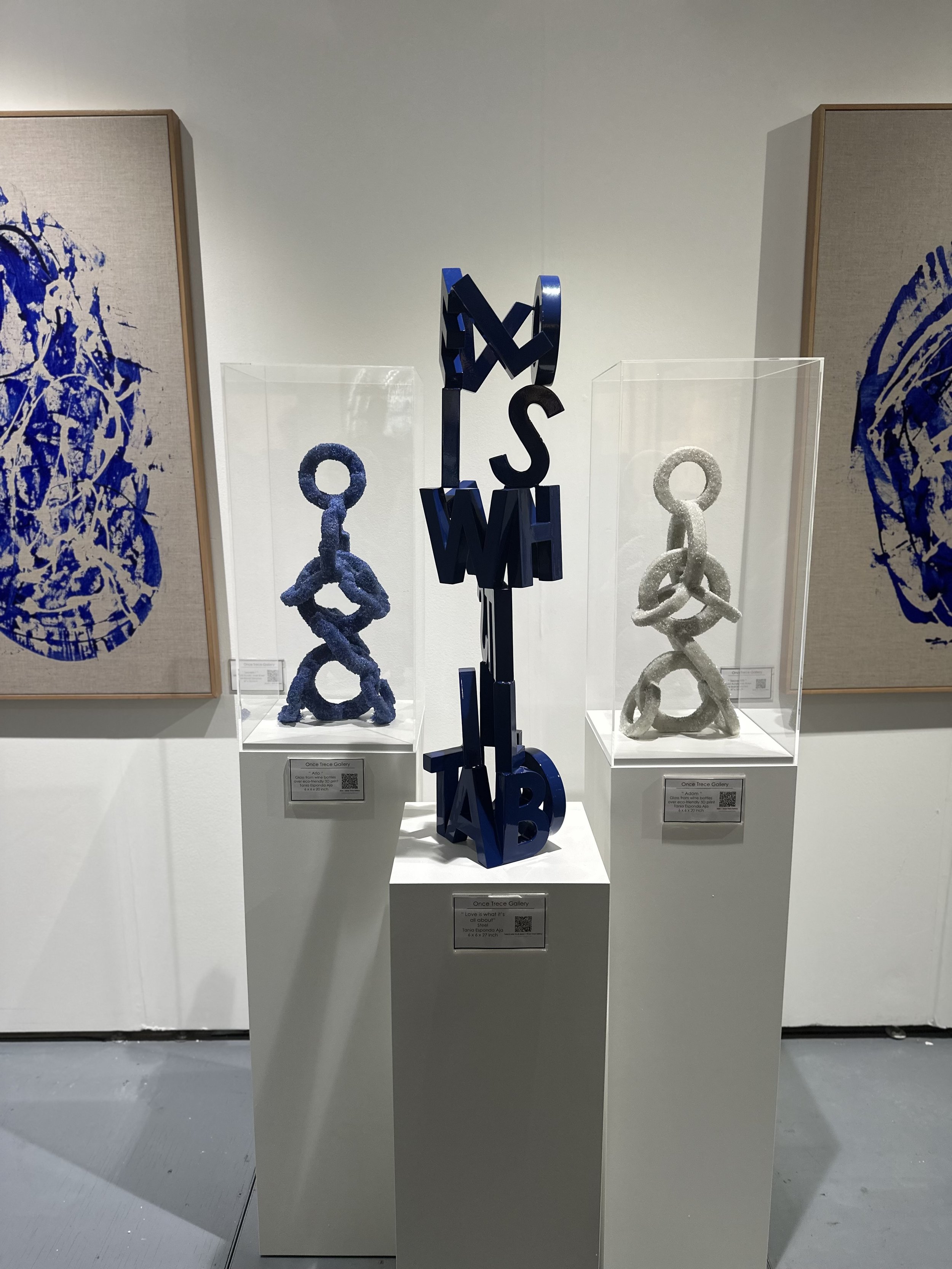 Contemporary art exhibit featuring blue and silver sculptures on pedestals, with abstract blue paintings on beige canvas in the background.
