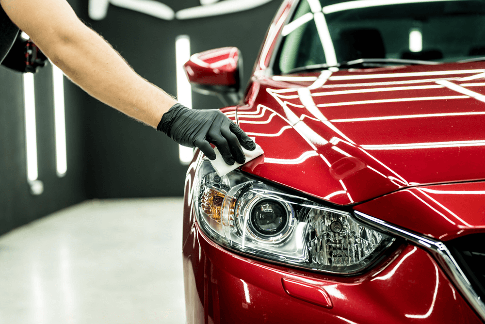 Person wearing black gloves polishing a red car's headlight with a cloth in a car detailing shop.