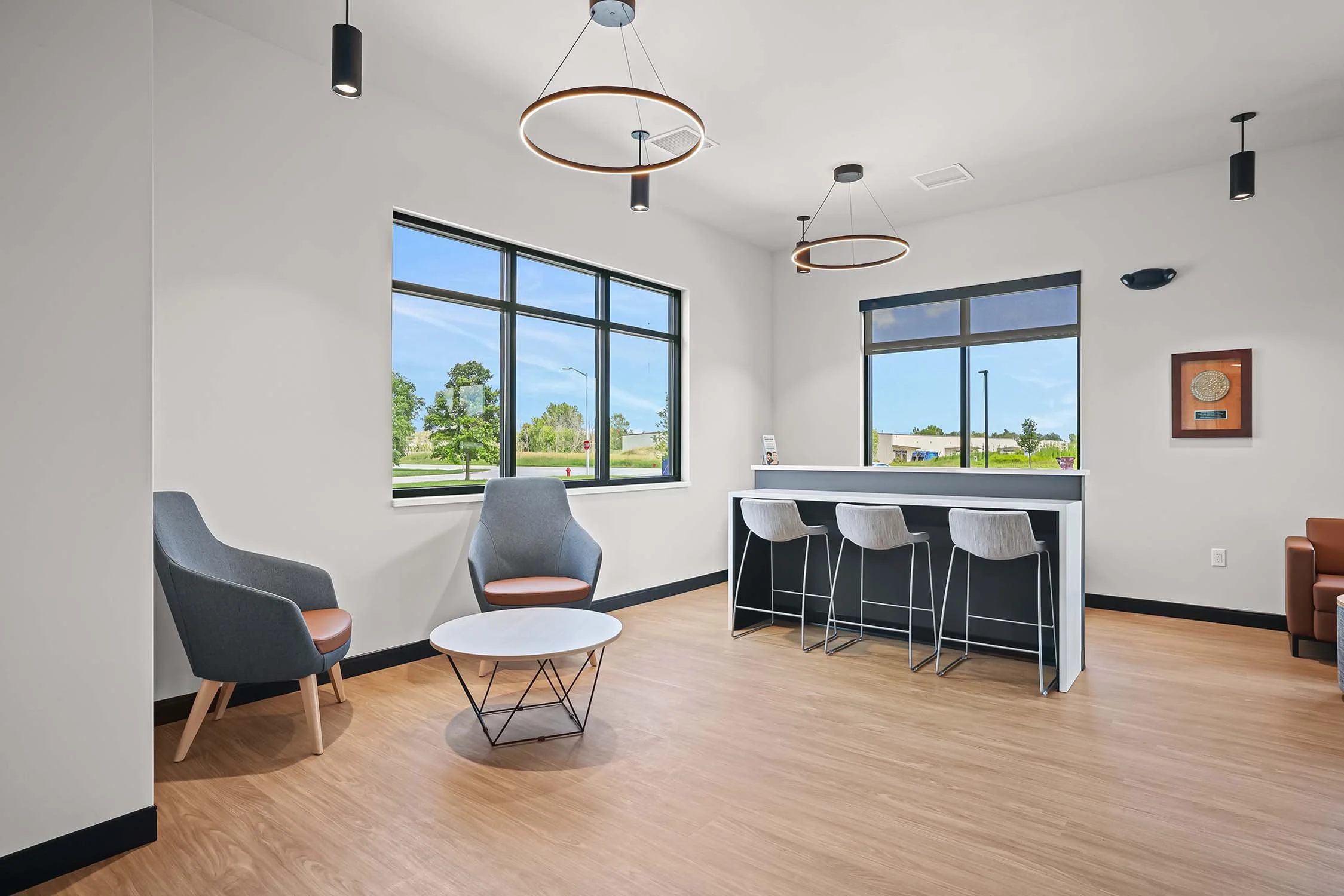 A modern, clean waiting room with beige walls, light wood flooring, large windows, 2 gray armchairs with brown cushions, a white round coffee table, a formica bar-height table with three gray bar stools, and contemporary lighting fixtures.