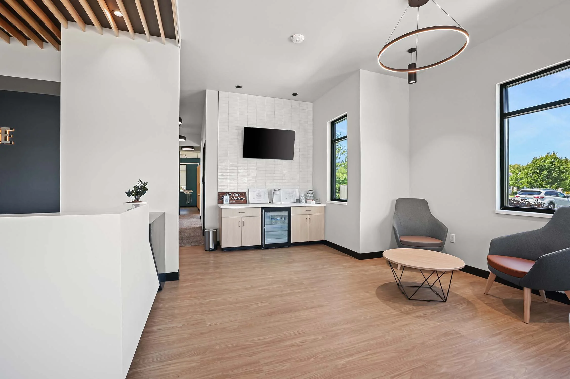 Modern waiting area with two gray chairs with brown cushions, a small round wooden table, large windows showing a parking lot and trees outside, and a cabinet with a TV mounted above it.