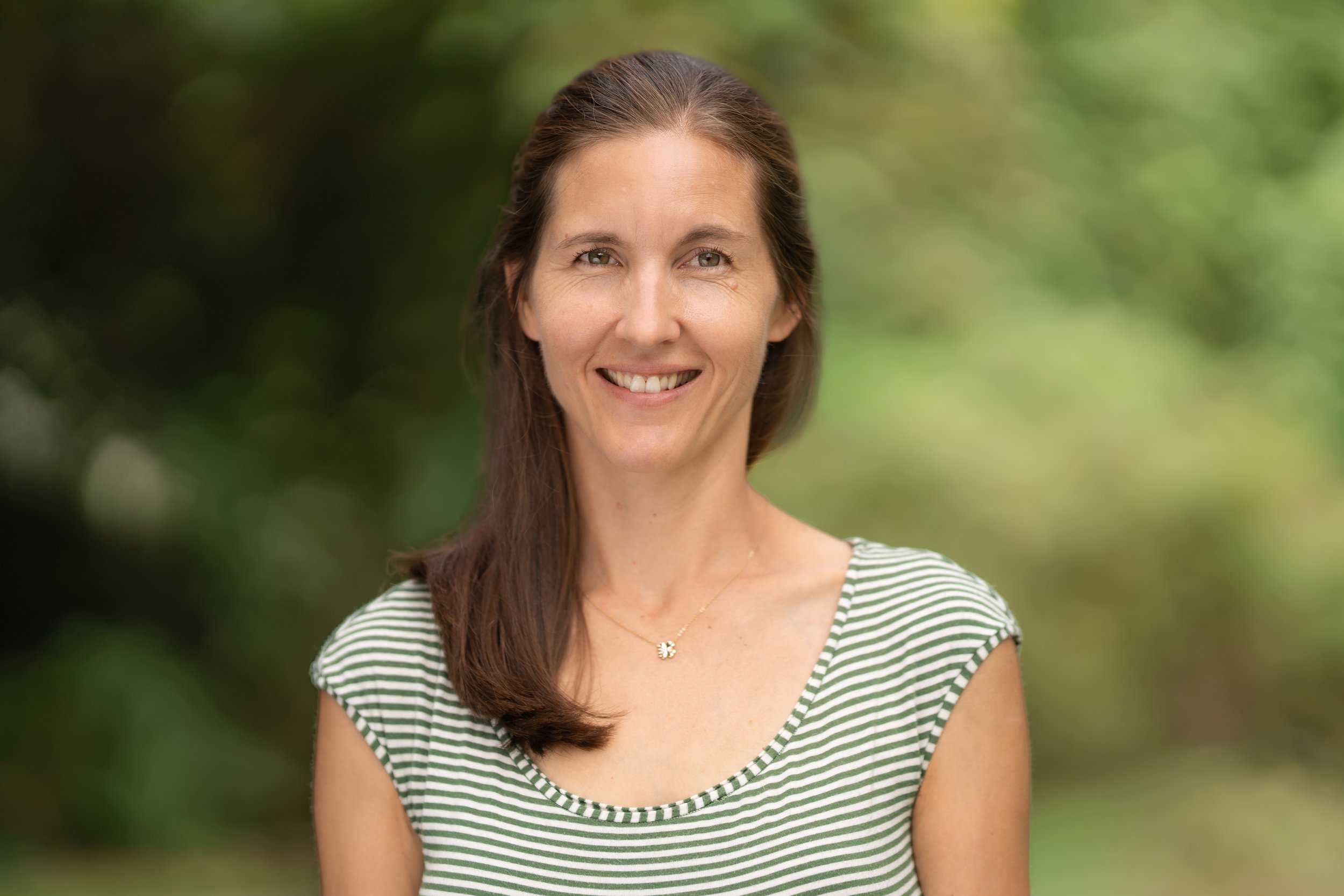 A smiling Grades Waldorf teacher standing outdoors with green foliage in the background.