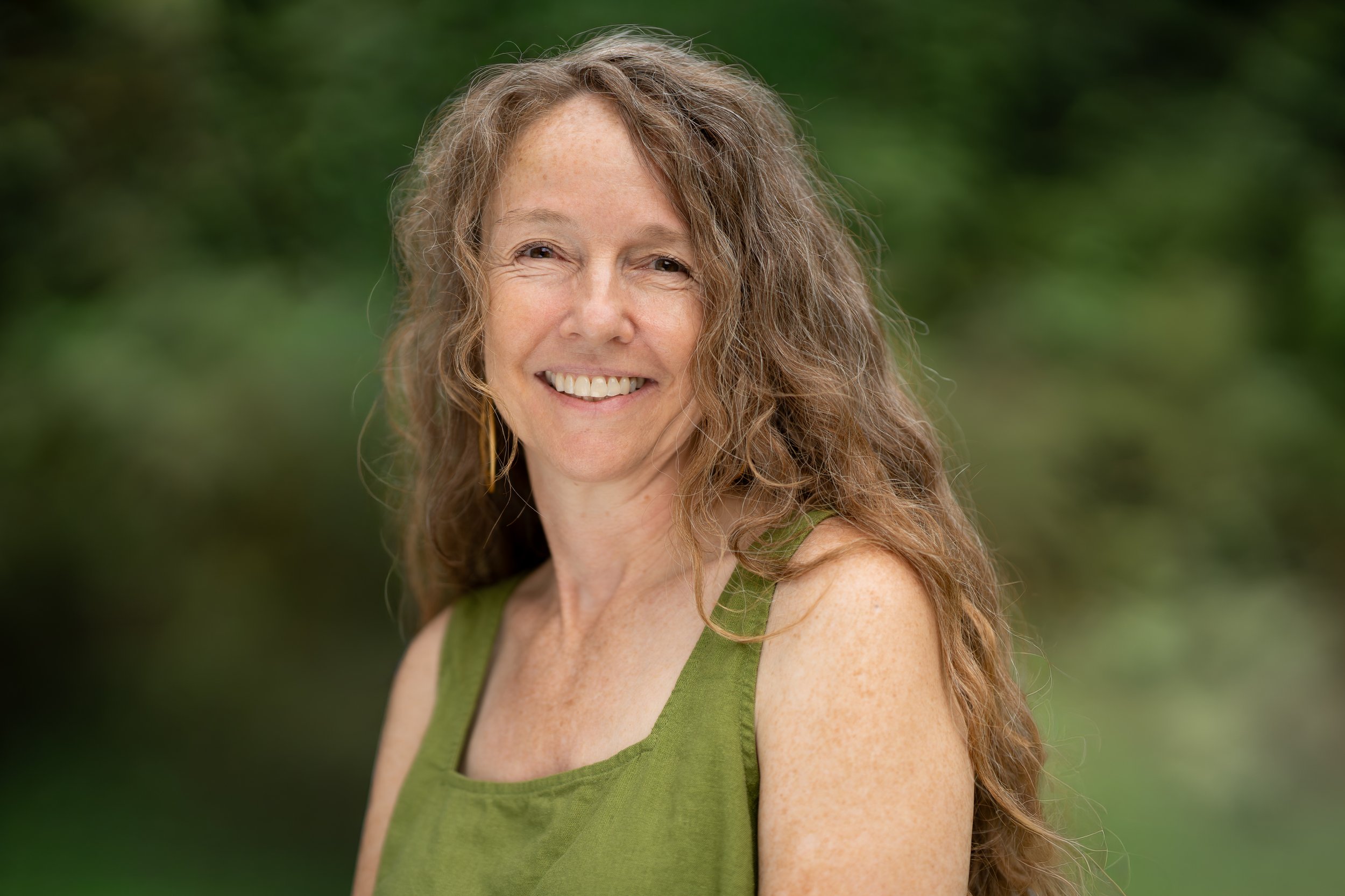 A smiling Waldorf teacher standing outdoors with green foliage in the background.