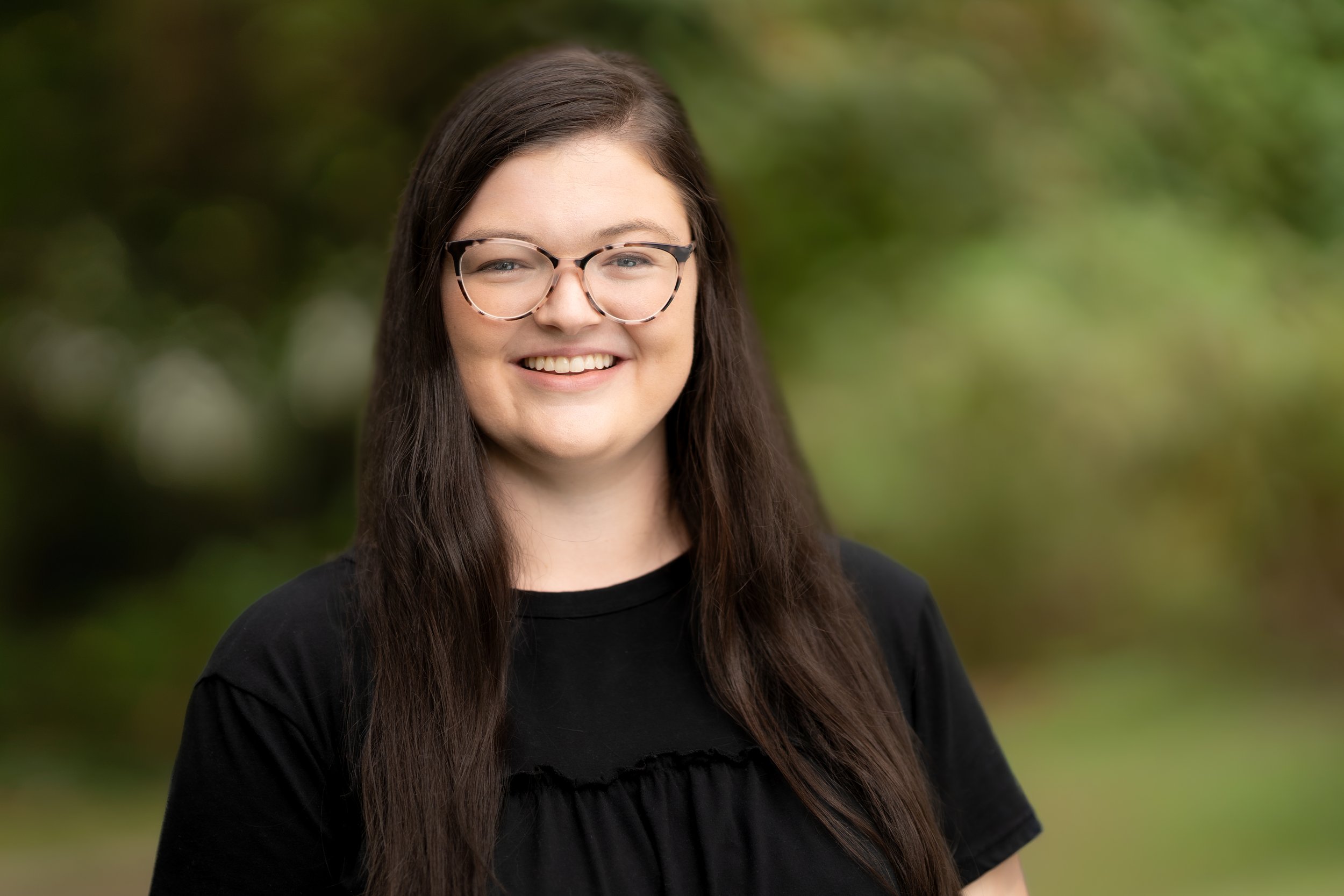 A smiling Music Waldorf teacher standing outdoors with green foliage in the background