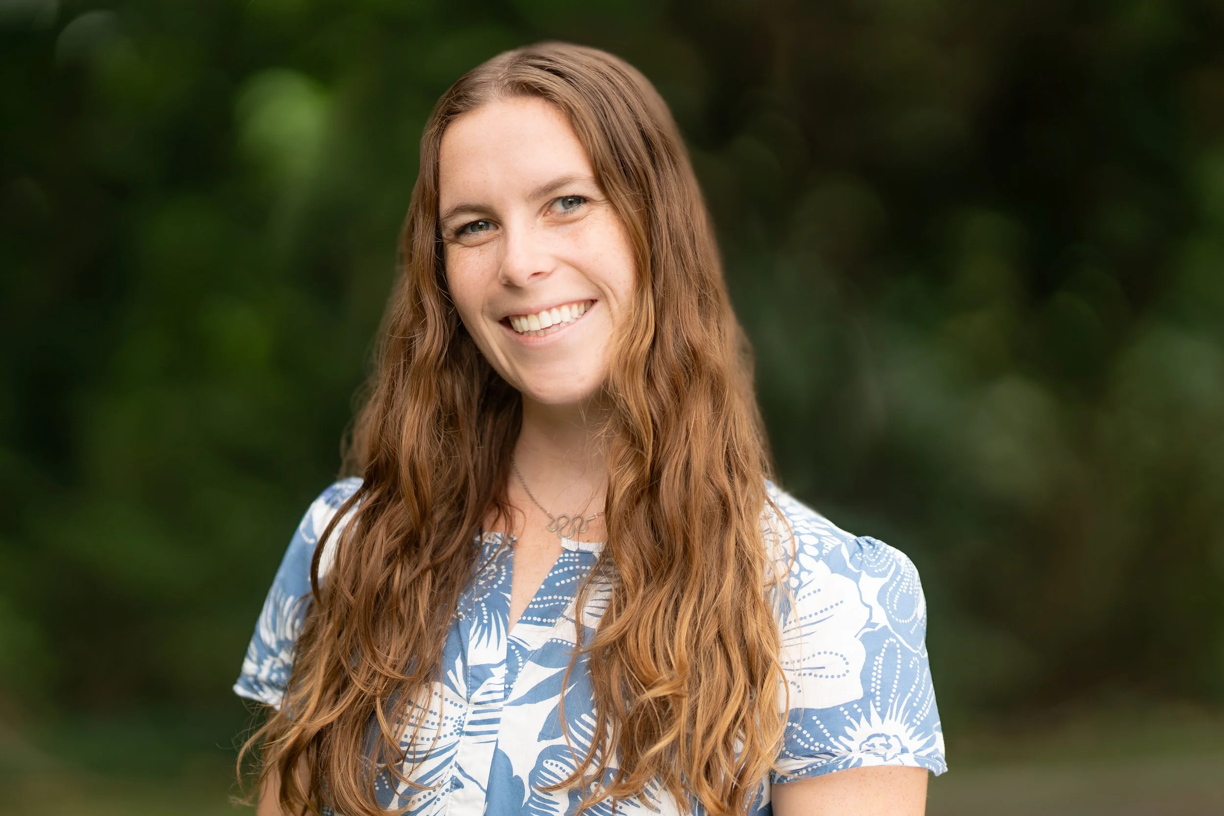 A smiling Grades Waldorf teacher standing outdoors with green foliage in the background.