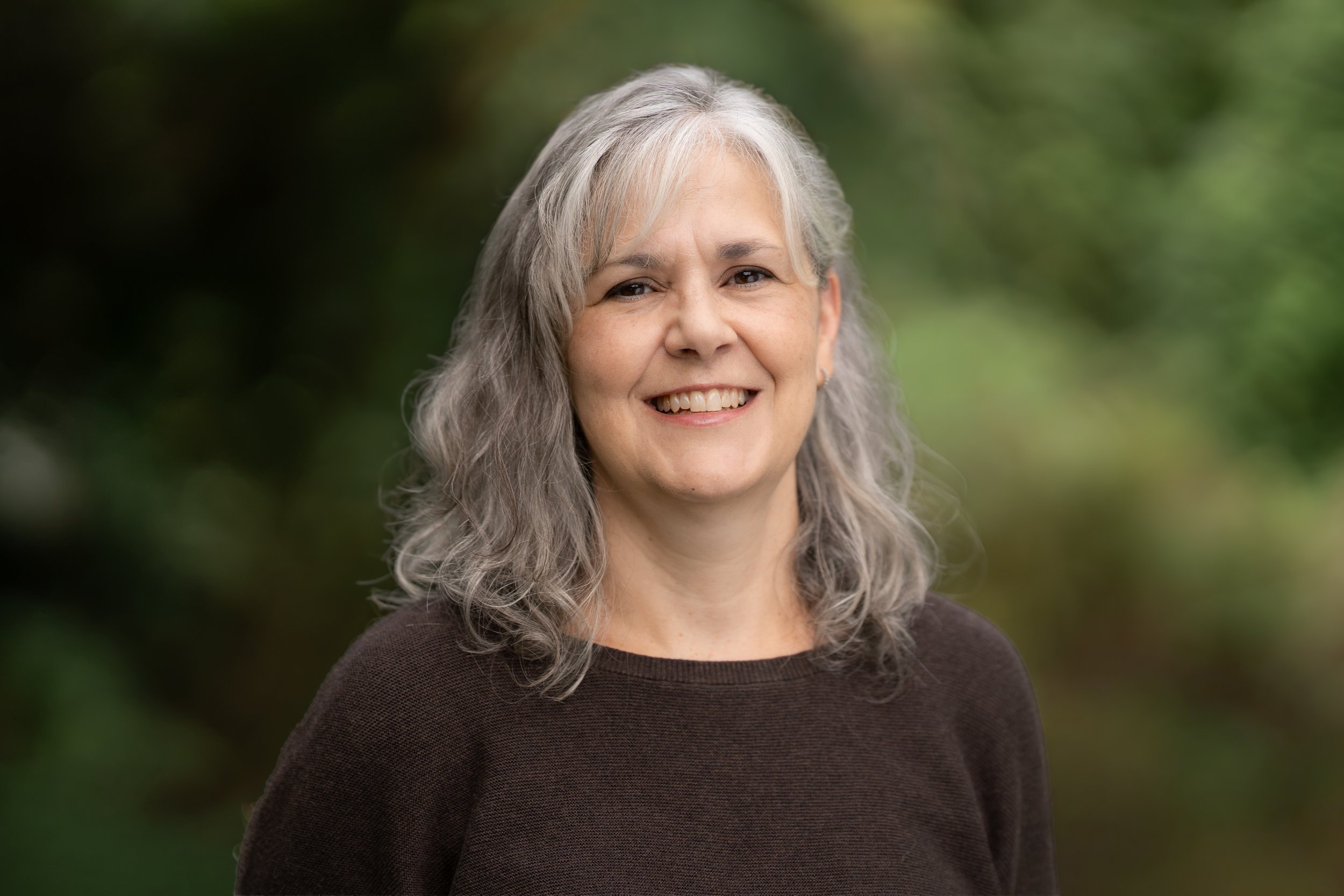 A smiling Forest Kindergarten Waldorf teacher standing outdoors with green foliage in the background.