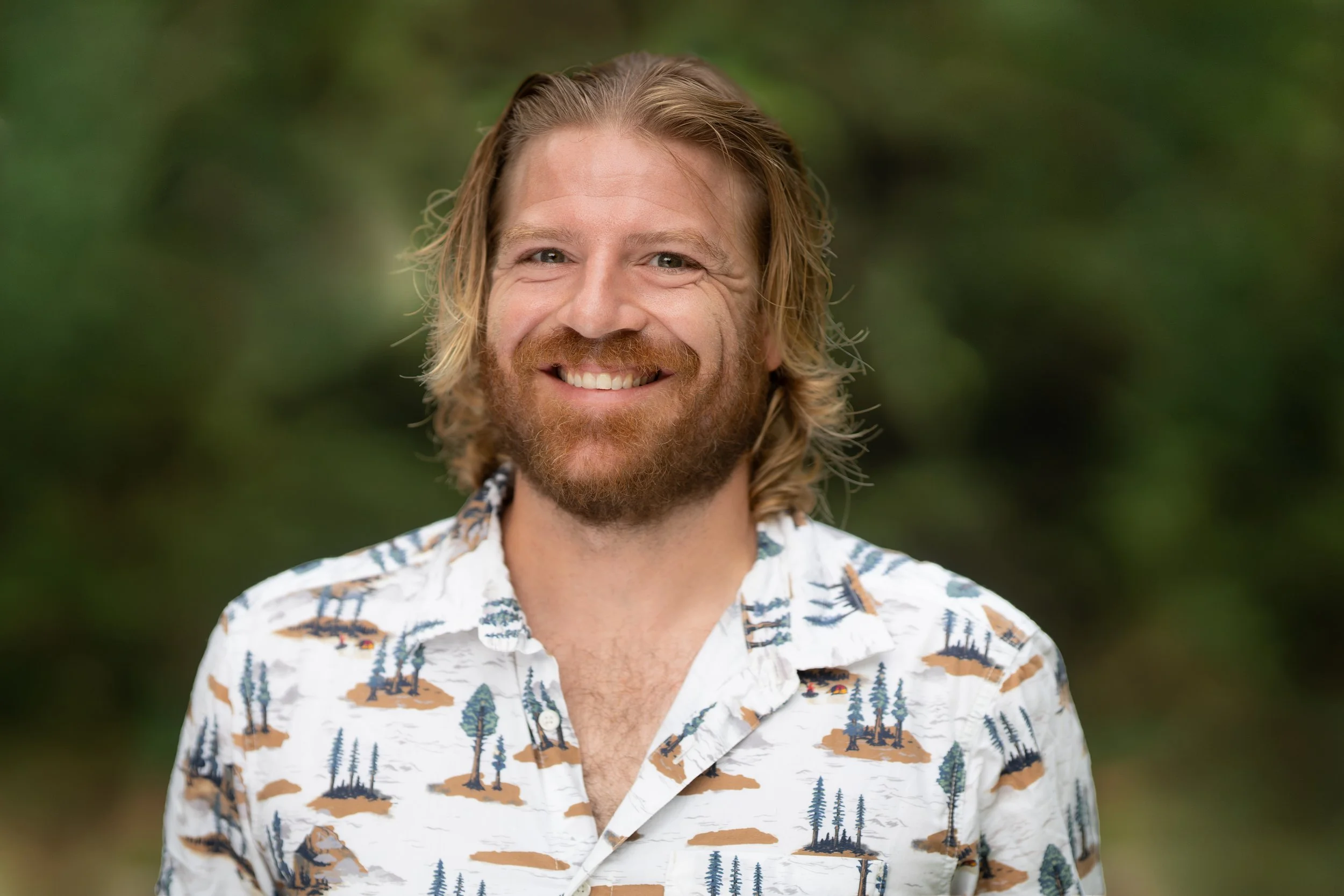 A smiling  Waldorf teacher standing outdoors with green foliage in the background.