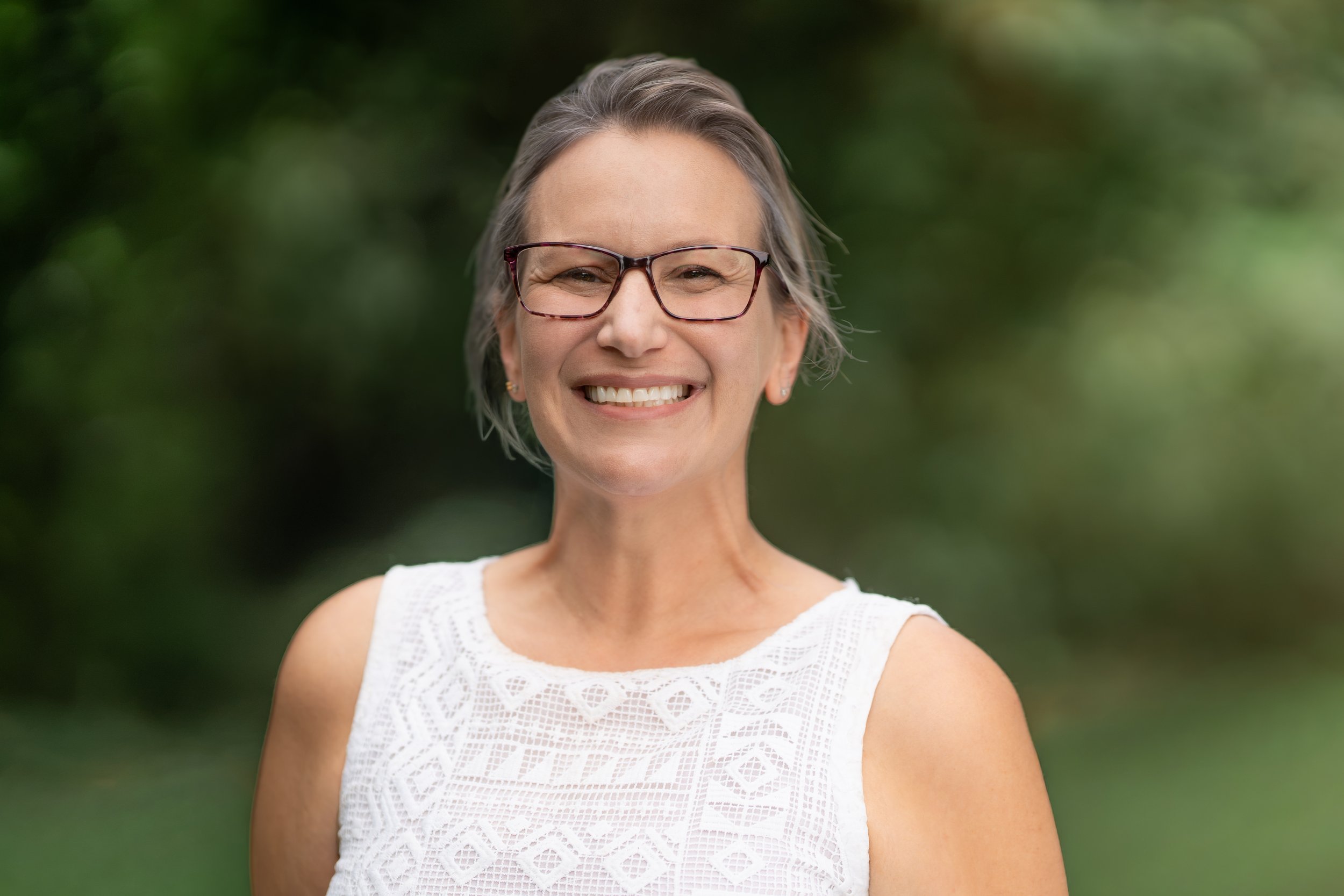 A smiling Forest Kindergarten Waldorf teacher standing outdoors with green foliage in the background.