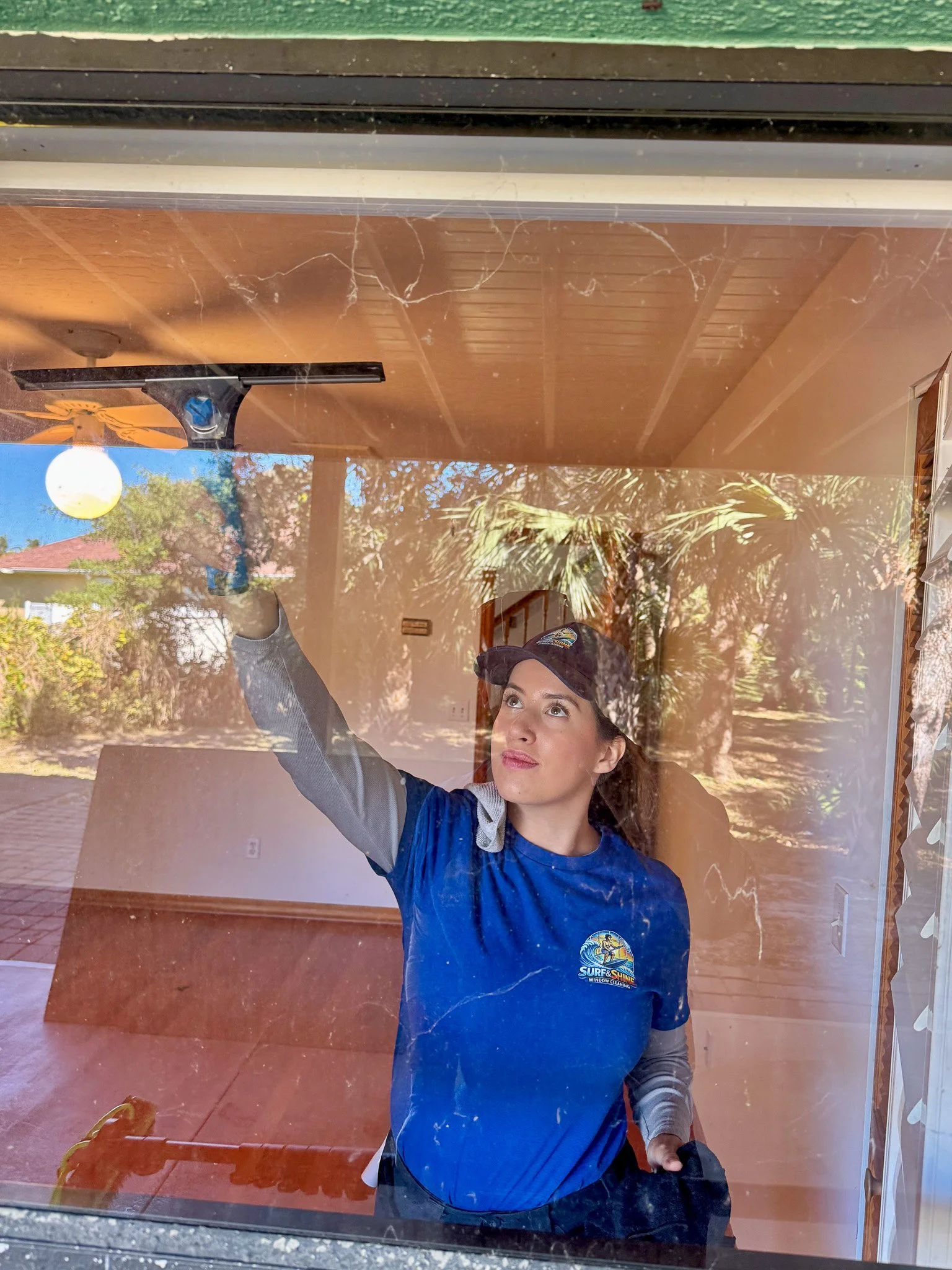 A woman cleaning a window from the inside, wearing a blue shirt and a dark cap, with reflections of trees and a house outside.