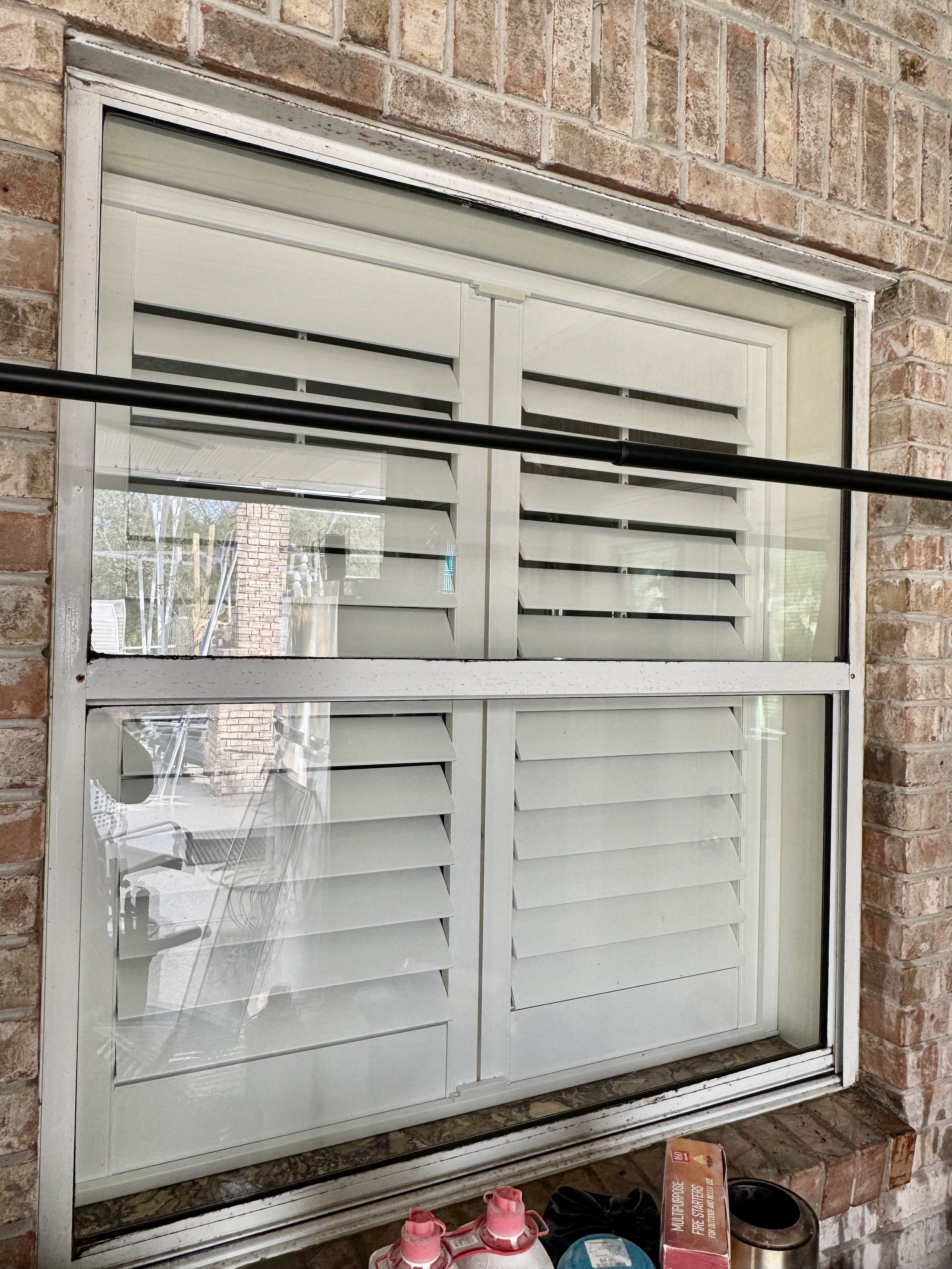 A window with white shutters in a brick wall, reflecting outdoor scenery. A black rod crosses the window, and some objects including pink shoes and cleaning supplies are at the bottom.