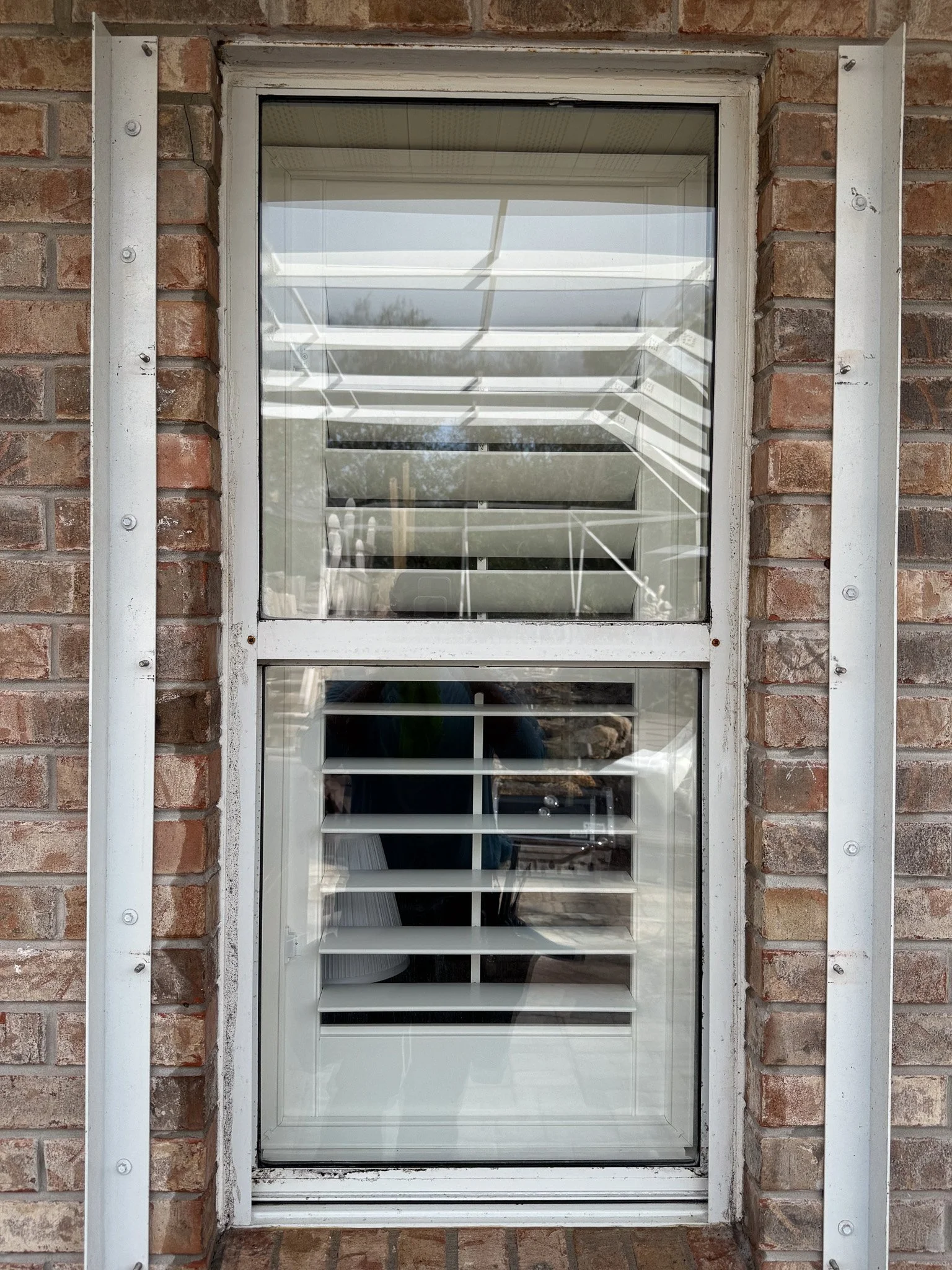 A brick wall window with white framing and horizontal blinds, reflecting part of a porch or patio with a white railing and some outdoor furniture.