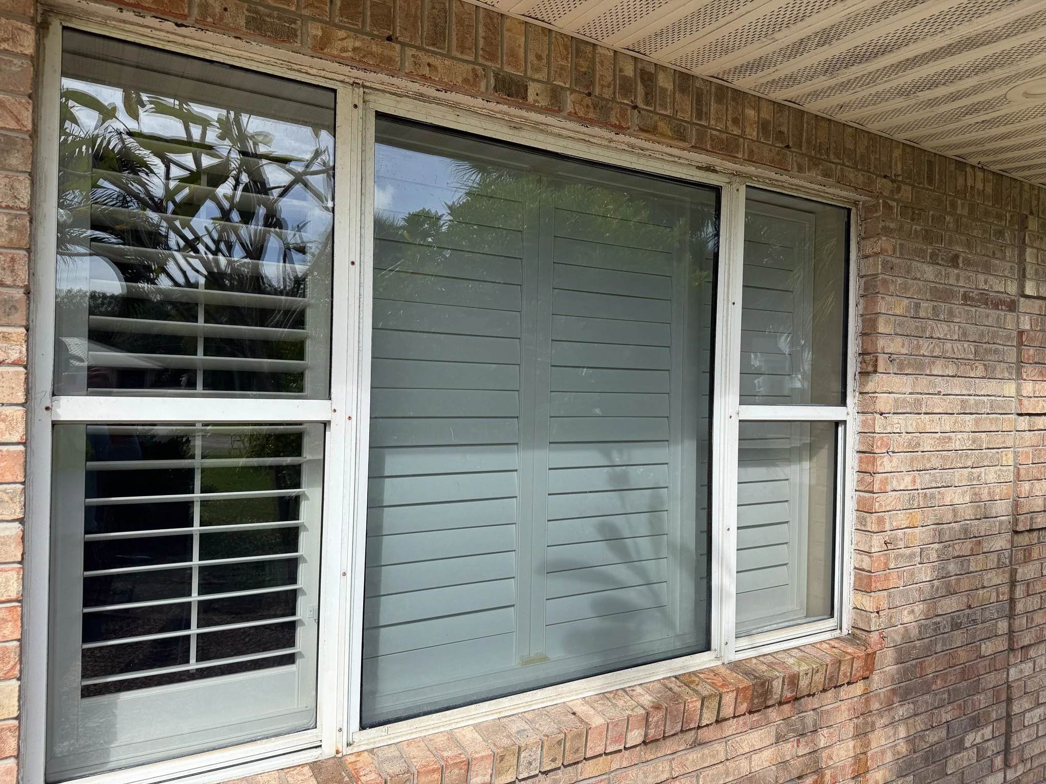 A brick house exterior window with white frame and horizontal blinds, partially open shutter on the left with decorative louvered slats, and a brick ledge underneath.