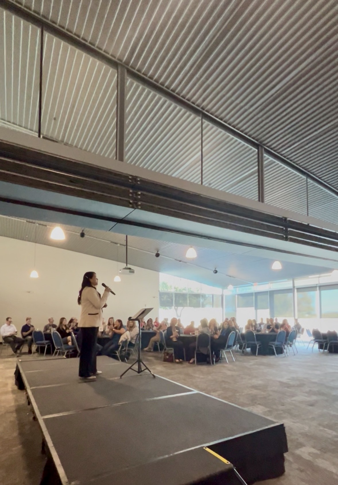 A woman speaking into a microphone on a small stage during a conference or seminar in a large, well-lit room with an audience seated at round tables.