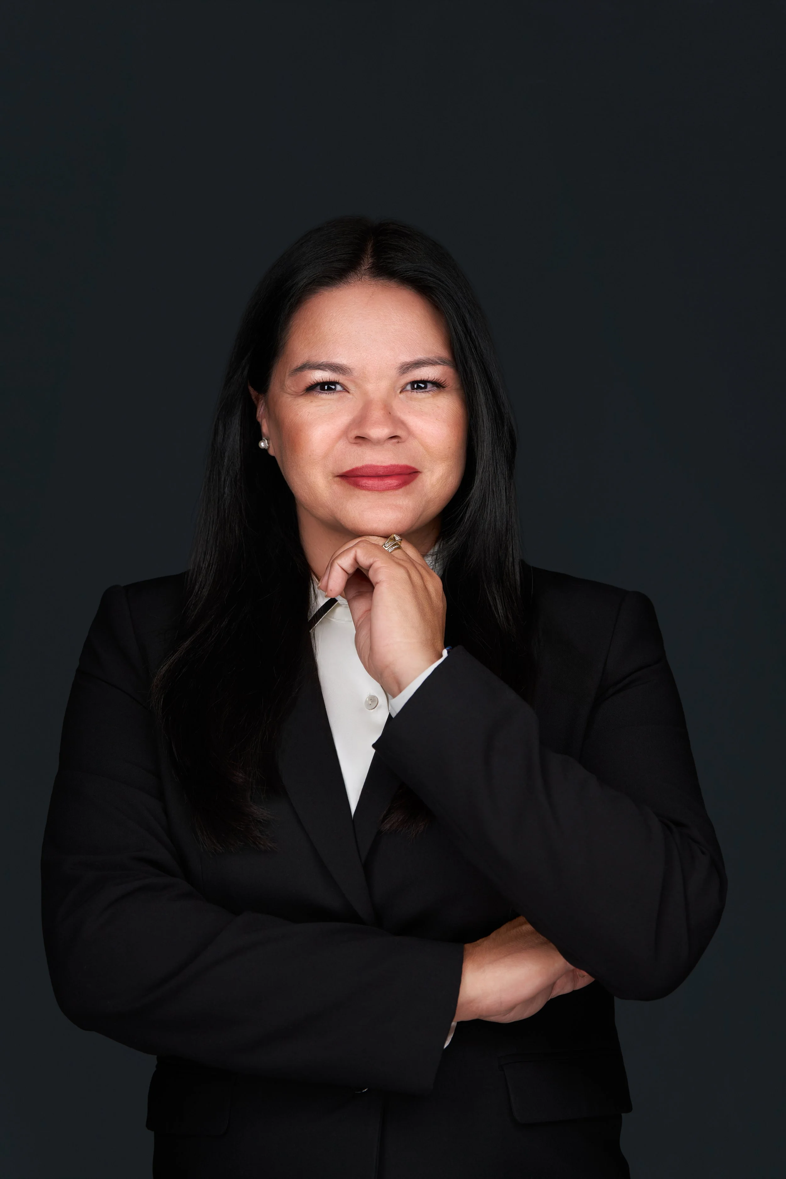 Professional woman with long black hair in a black blazer, white shirt, standing against a dark background, looking confidently at the camera with her hand resting on her chin.