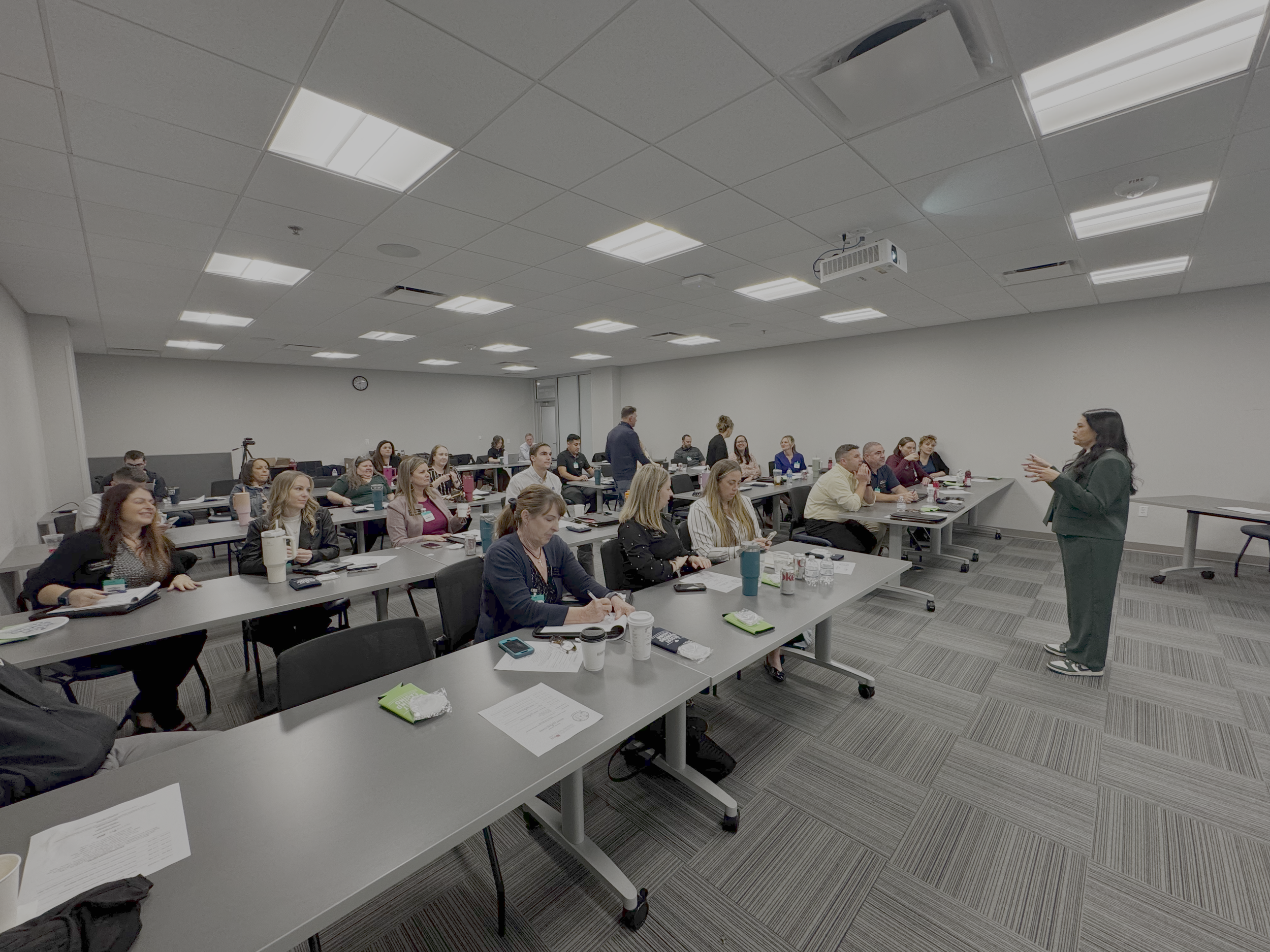 A woman with black hair, dressed in a dark blazer and pants, is speaking and gesturing with her hands to an audience of approximately 25 people in a modern conference room with gray walls, tiled ceiling, and carpeted floor.