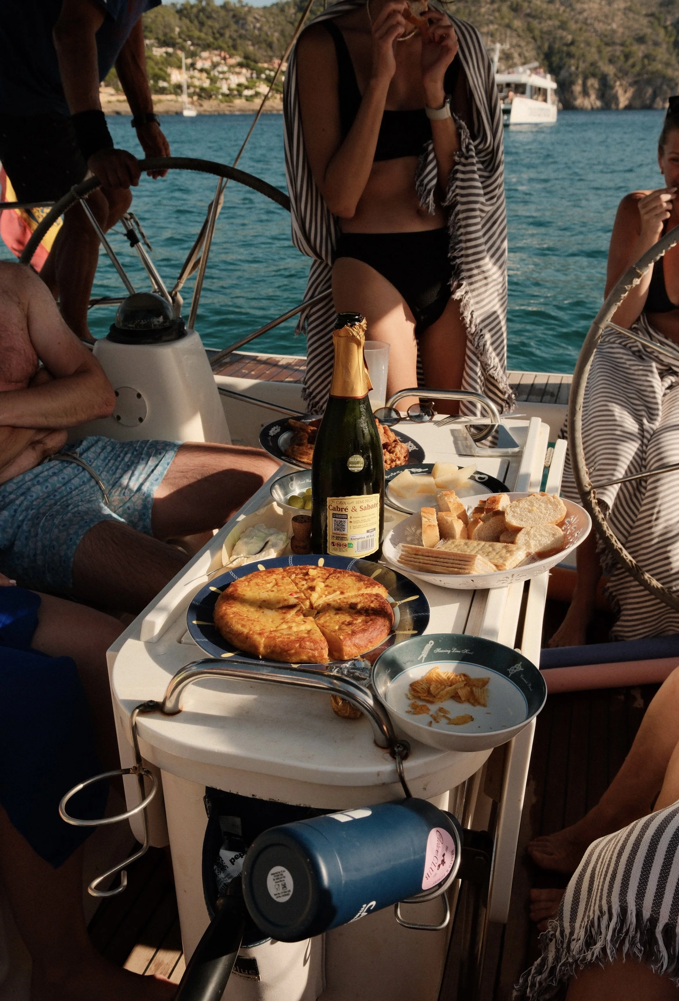 A group of people enjoying a meal on a boat with water and shoreline in the background. The table has pizza, bread, cheese, and a bottle of sparkling wine.