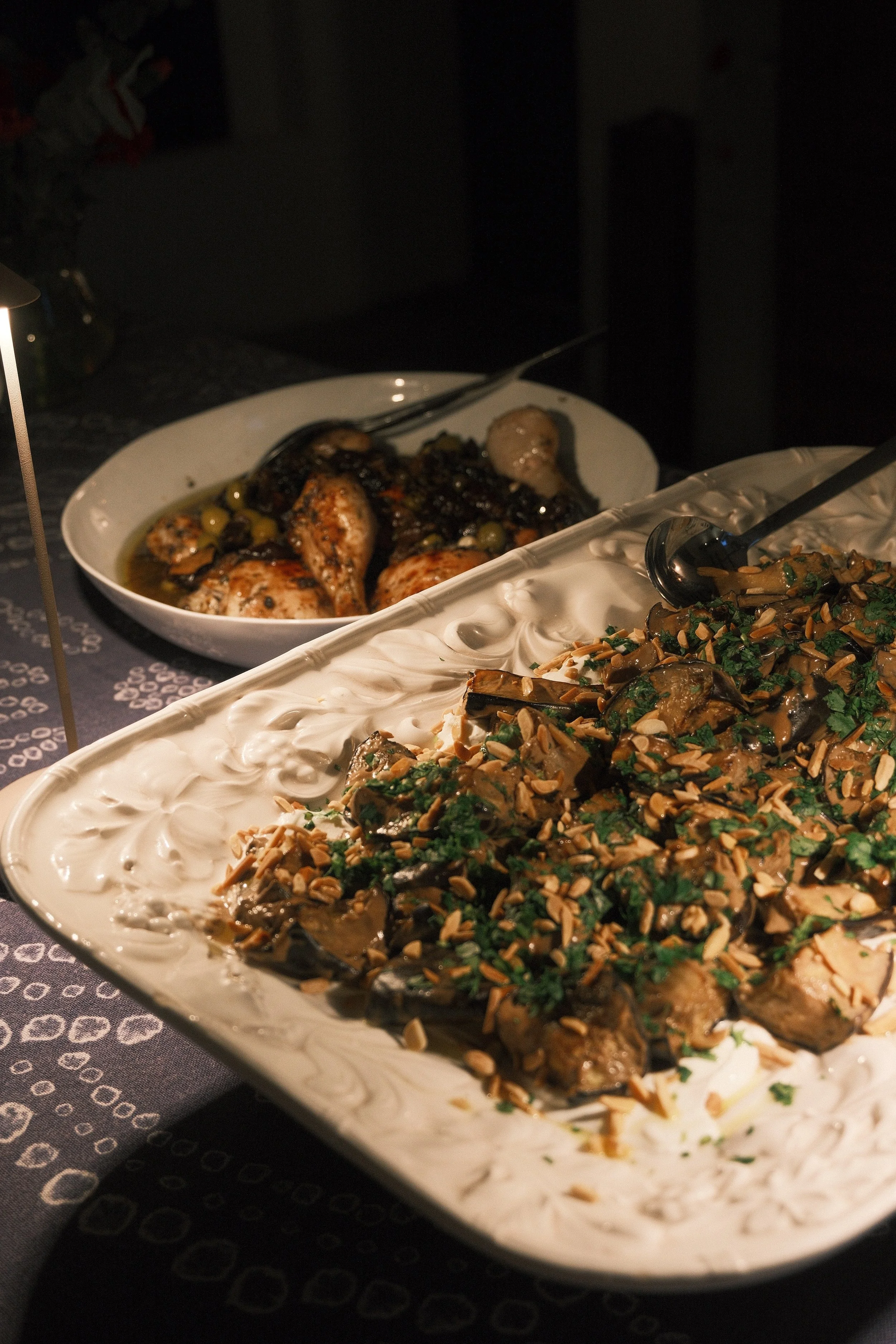 Close-up of a buffet table with dishes, including a white creamy dish topped with herbs and nuts, and a dish with cooked chicken with green olives in dark sauce in the background.