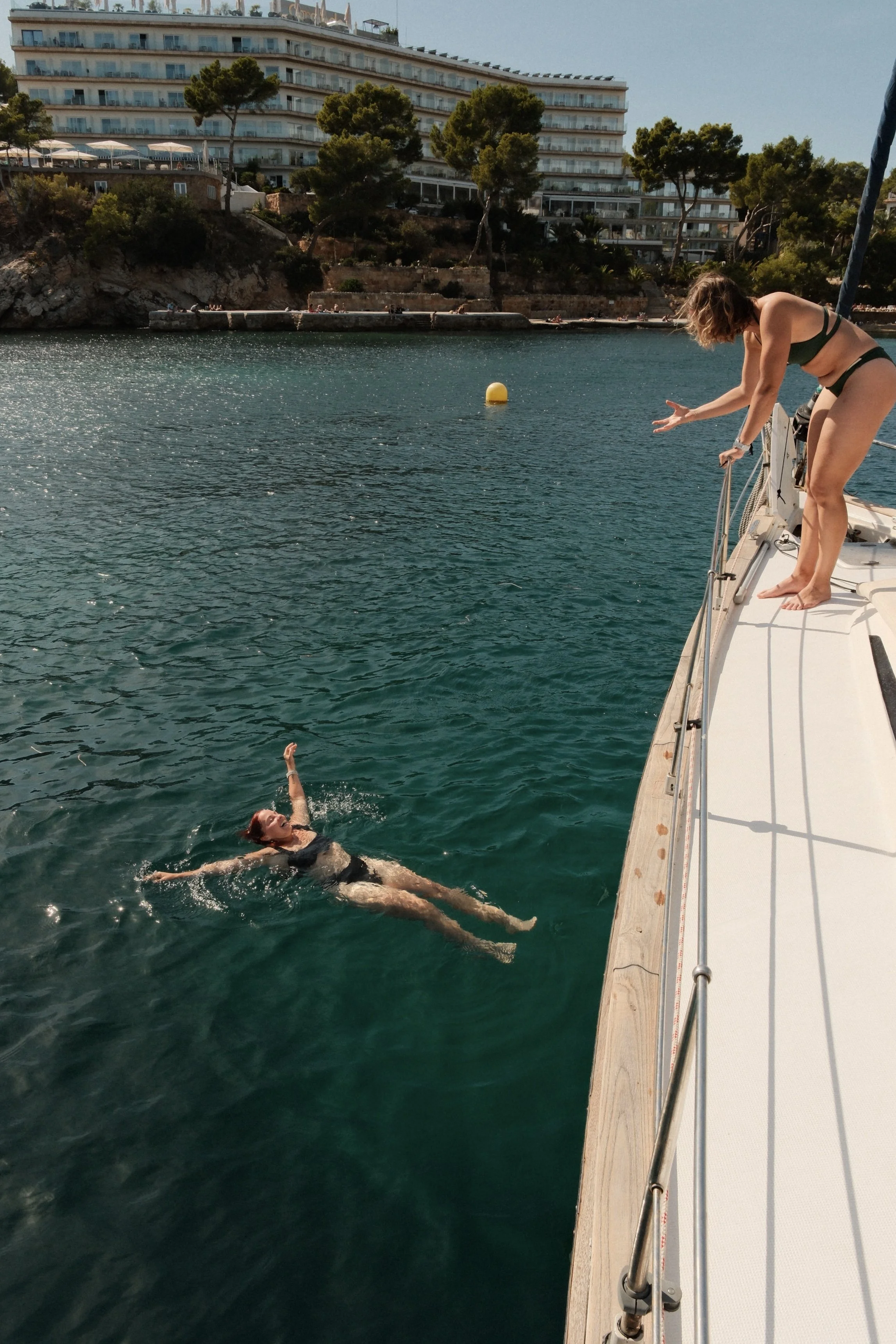A woman swimming in the water next to a boat, with another woman standing on the boat's deck reaching out to her. The scene includes a rocky shoreline, trees, and a large building in the background.