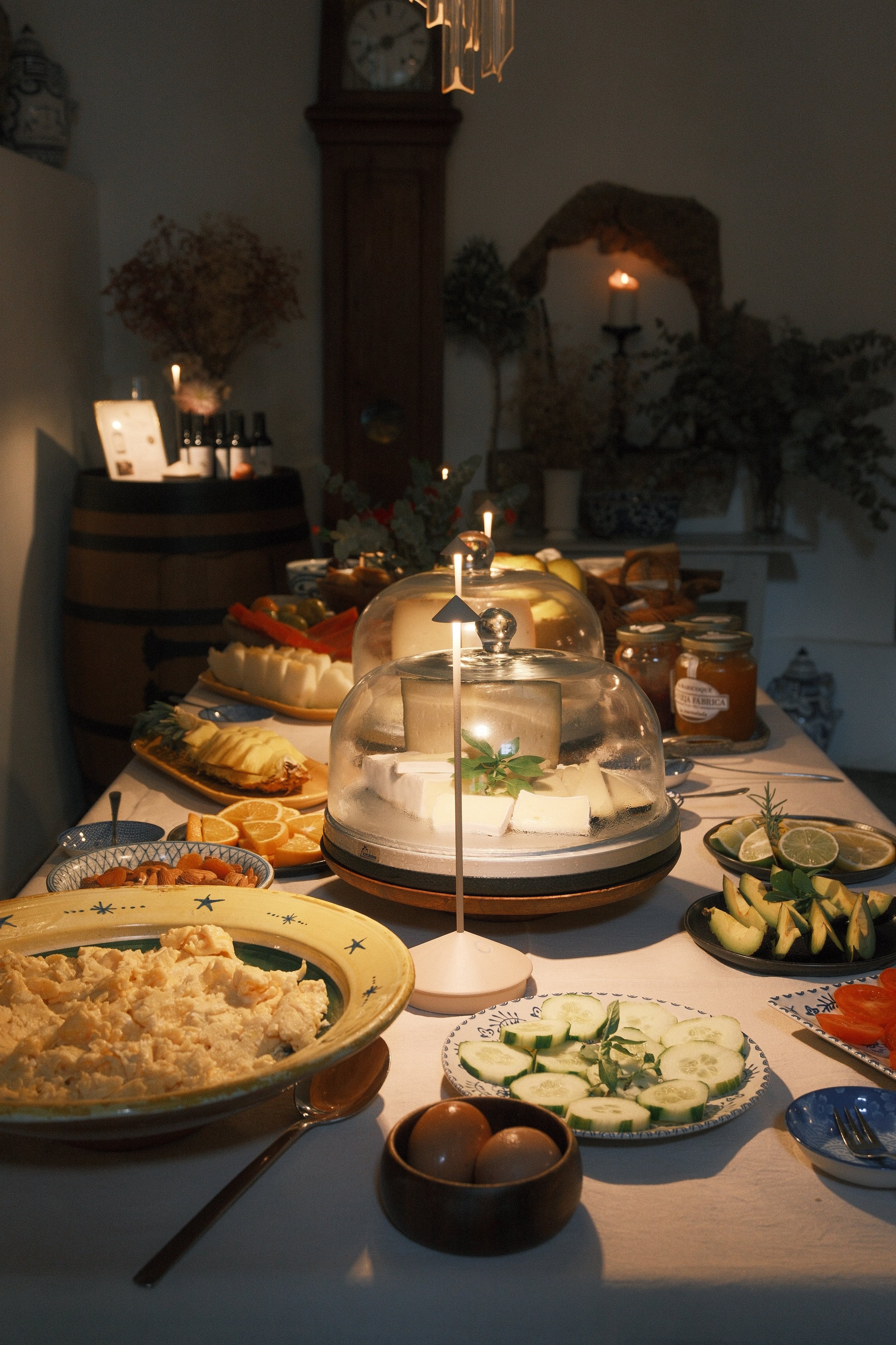 A dinner table with various cheeses, sliced vegetables, and dips, illuminated by candles and a glass cake dome in a cozy room with dried flowers and vintage decor.