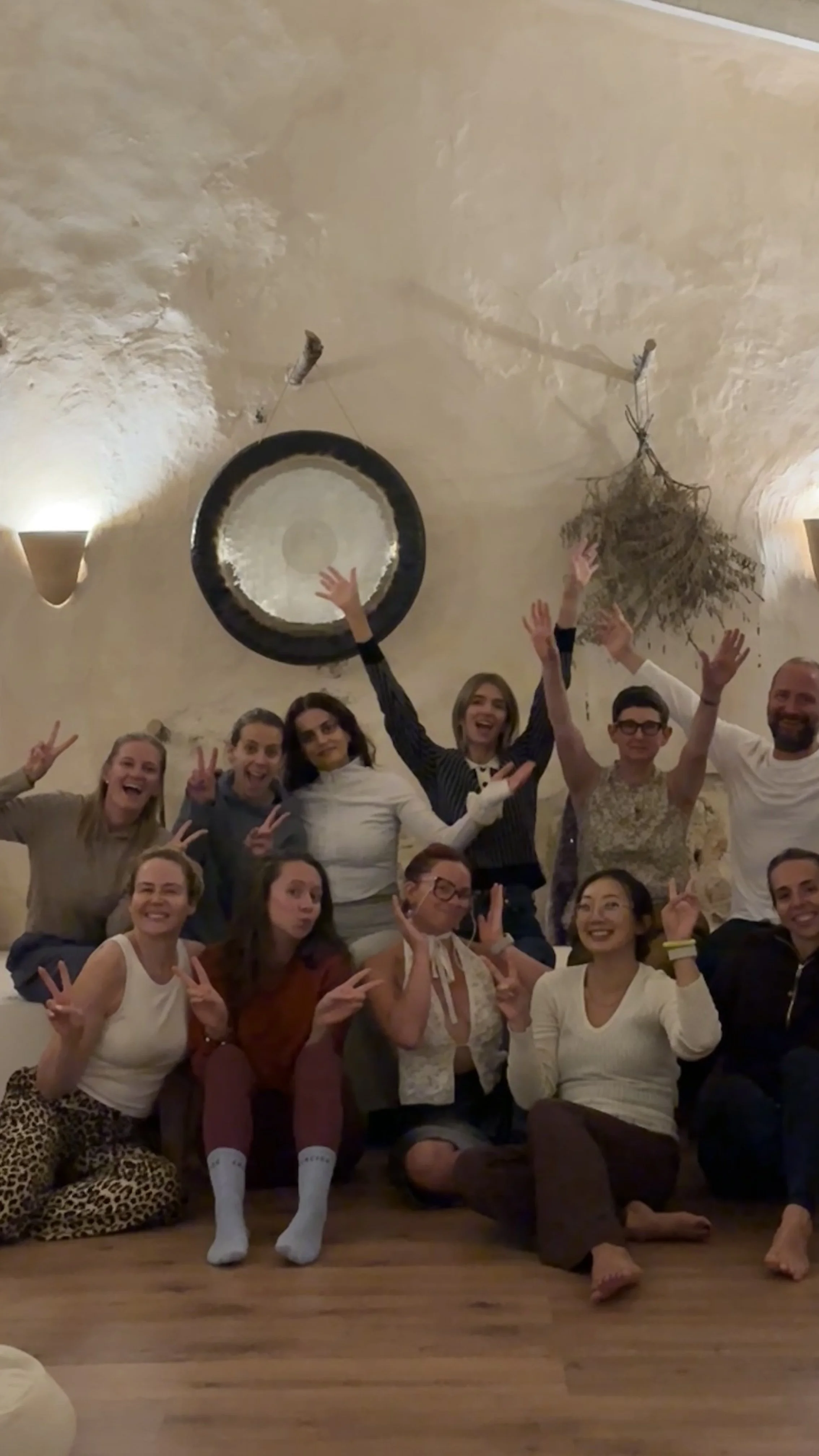 A group of people gathered in a room, posing happily for a photo. Some are sitting on the floor, others standing behind them, making peace signs and raising their hands. The ceiling has a large gong and drying herbs hanging from it.