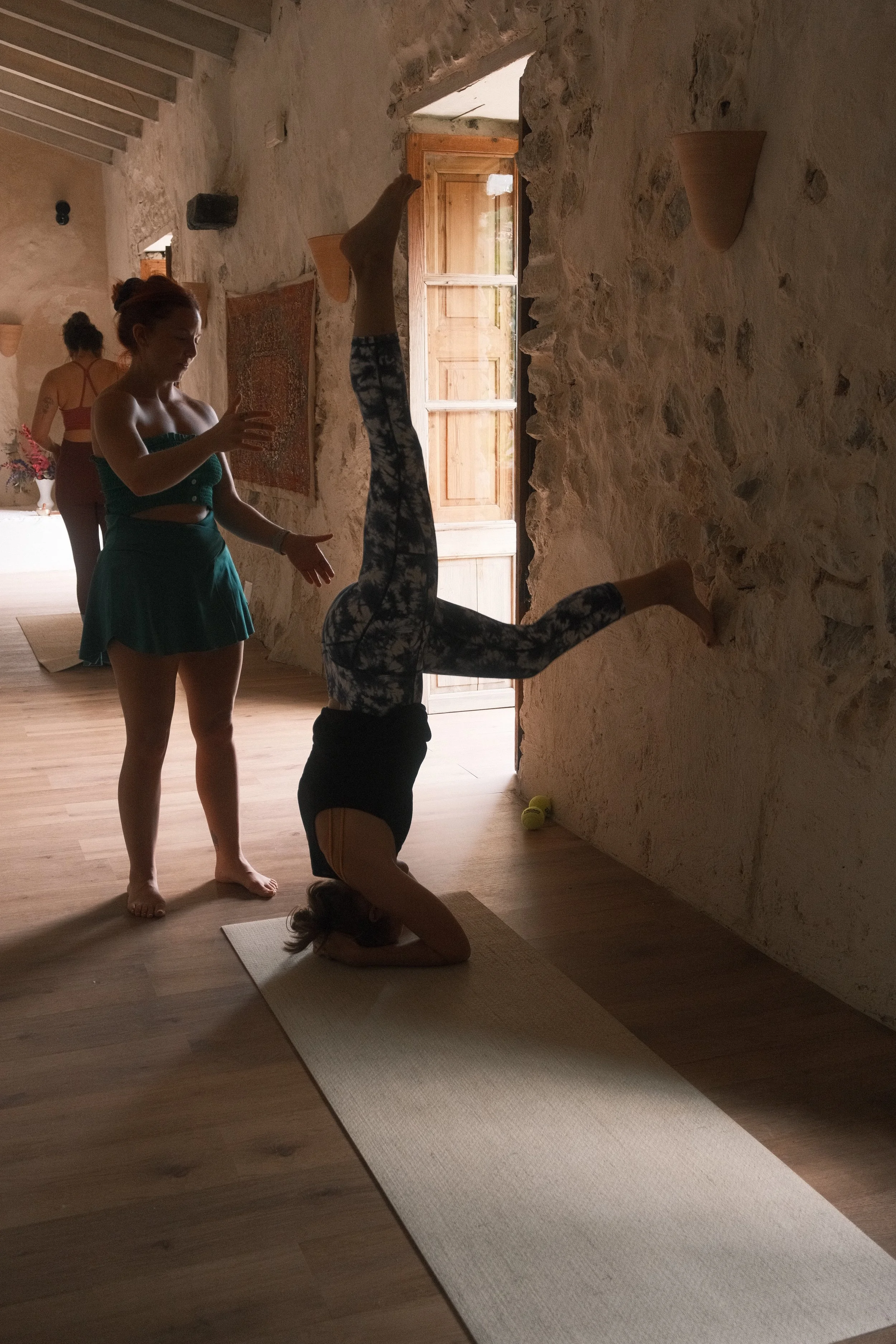 Two women practicing yoga indoors with stone walls. One is standing and watching, the other is in a headstand position on a yoga mat. Natural light coming through a wooden door.