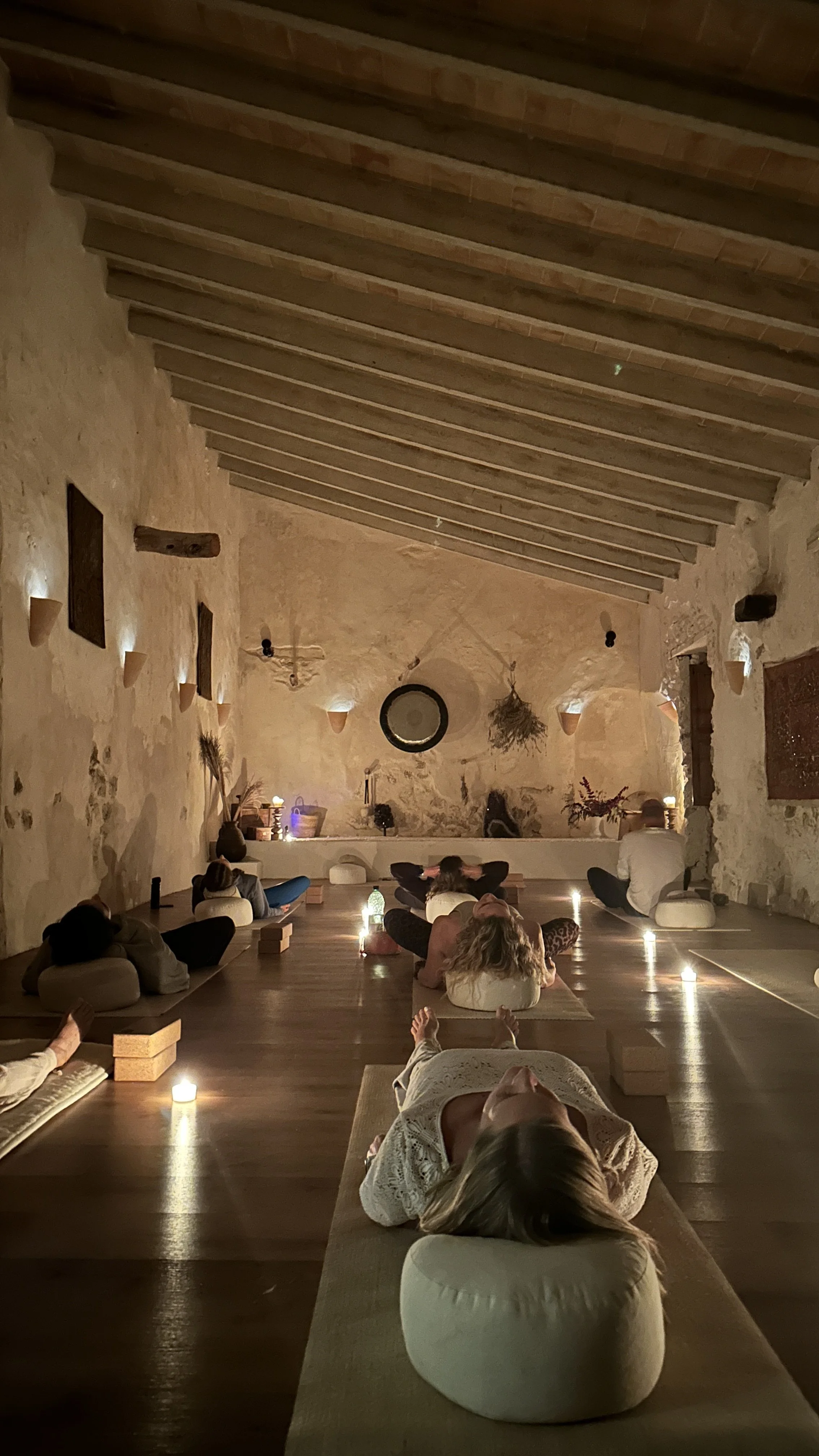 People sitting on cushions on the floor in a dimly lit room with candles, practicing meditation or yoga. The room has stone walls, wooden ceiling beams, and minimal decor, creating a peaceful atmosphere.