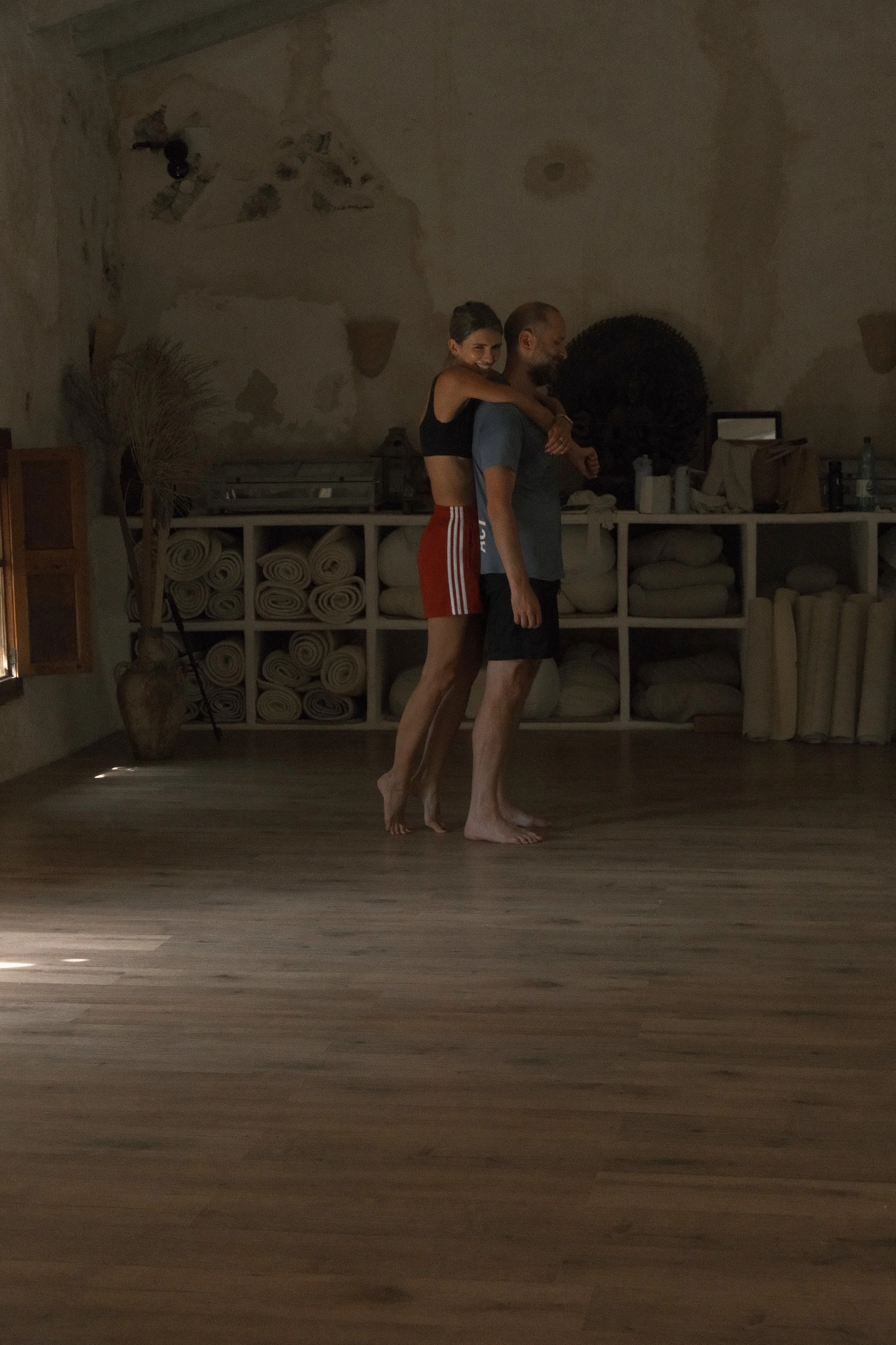 A woman and a man stand close together in a room with wooden flooring and a wall with visible patches. The woman is leaning on the man's back, smiling, while the man is standing with his eyes closed. The woman is wearing a black top and red striped s