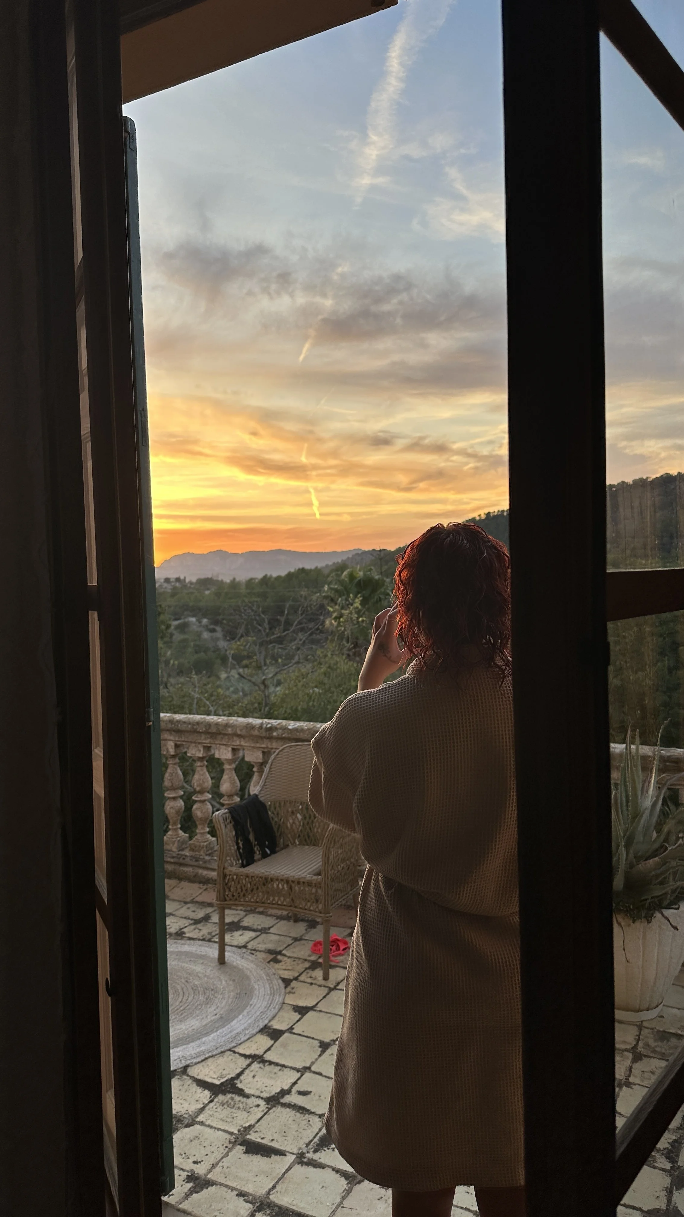 A woman with curly hair takes a picture of the sunset from a balcony, standing with her back to the camera, with trees and mountains in the background.