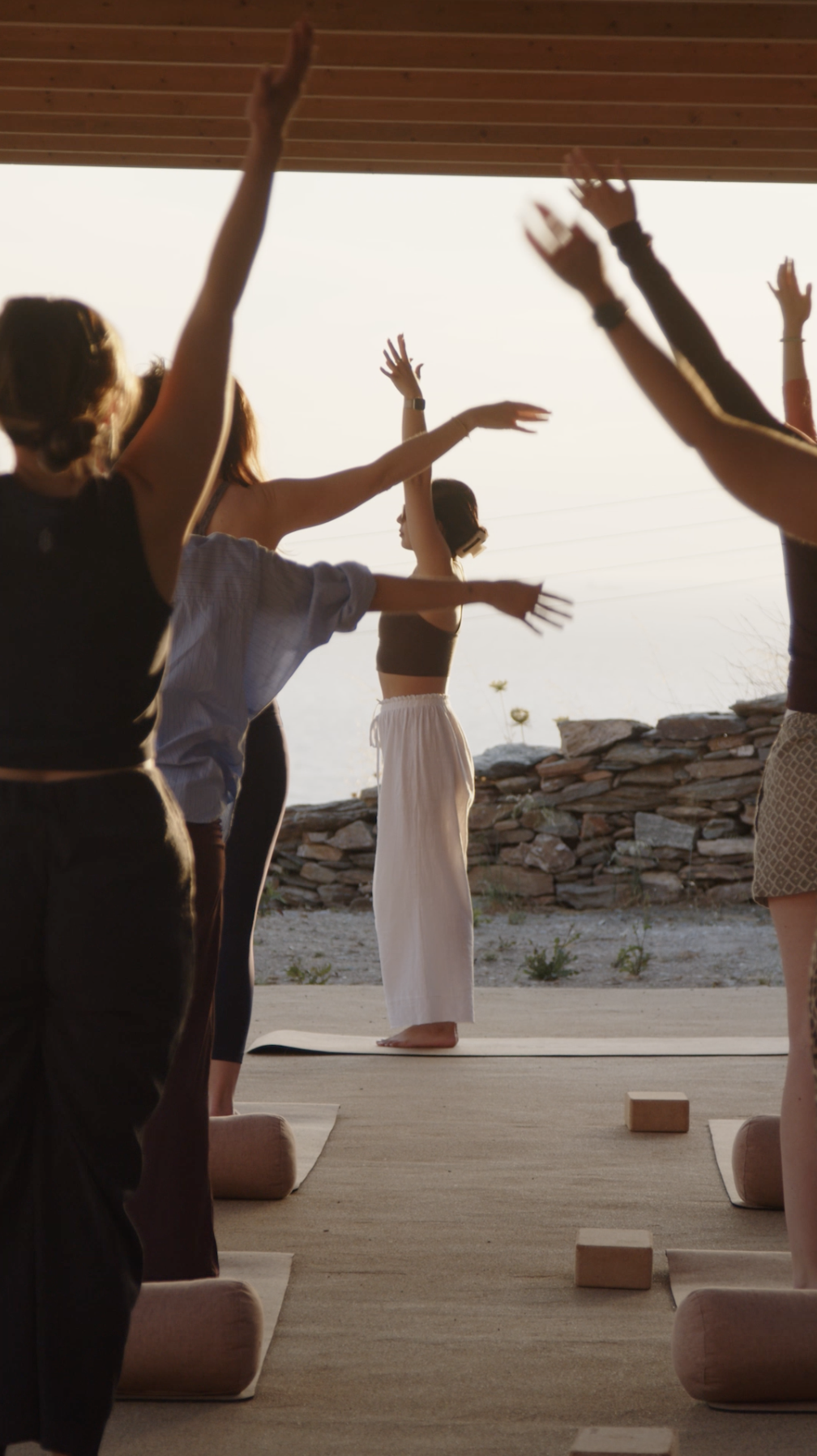 People participating in a yoga session outdoors during sunset, standing on yoga mats with their arms raised.