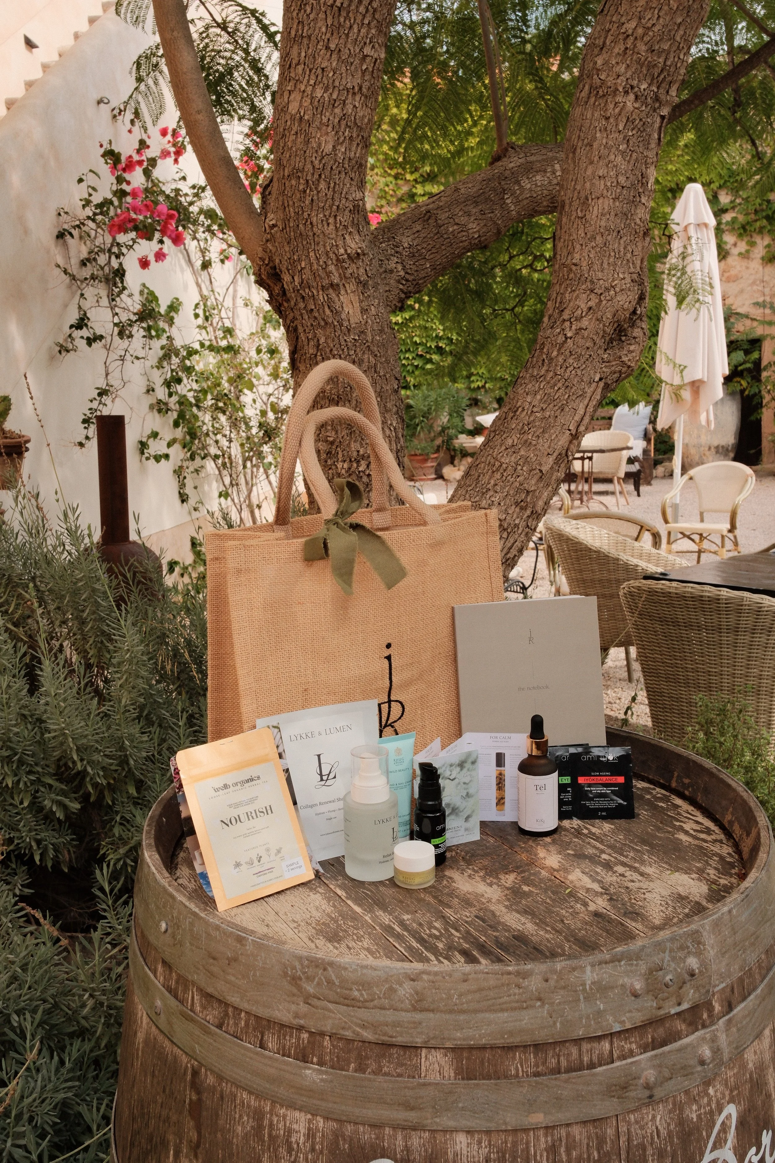 A wooden table displaying skincare and wellness products, with informational cards, set outdoors under a large tree with pink flowers and surrounded by chairs and umbrellas in the background.