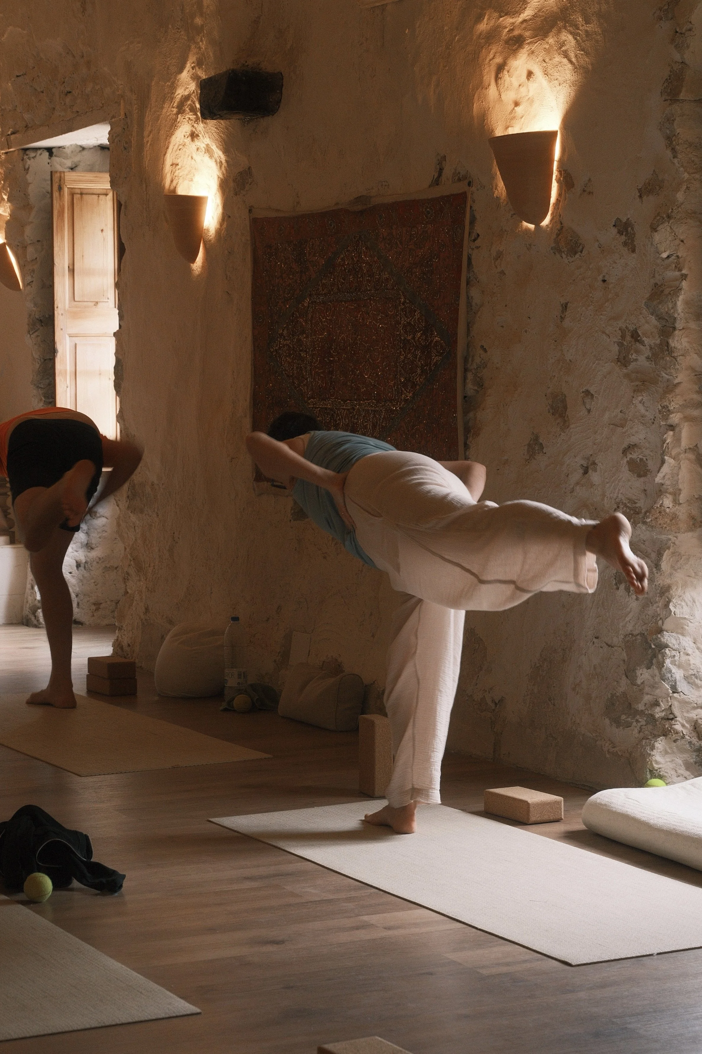 People practicing yoga in a room with stone walls, illuminated by warm wall lights, with yoga mats, blocks, and cushions on the floor.