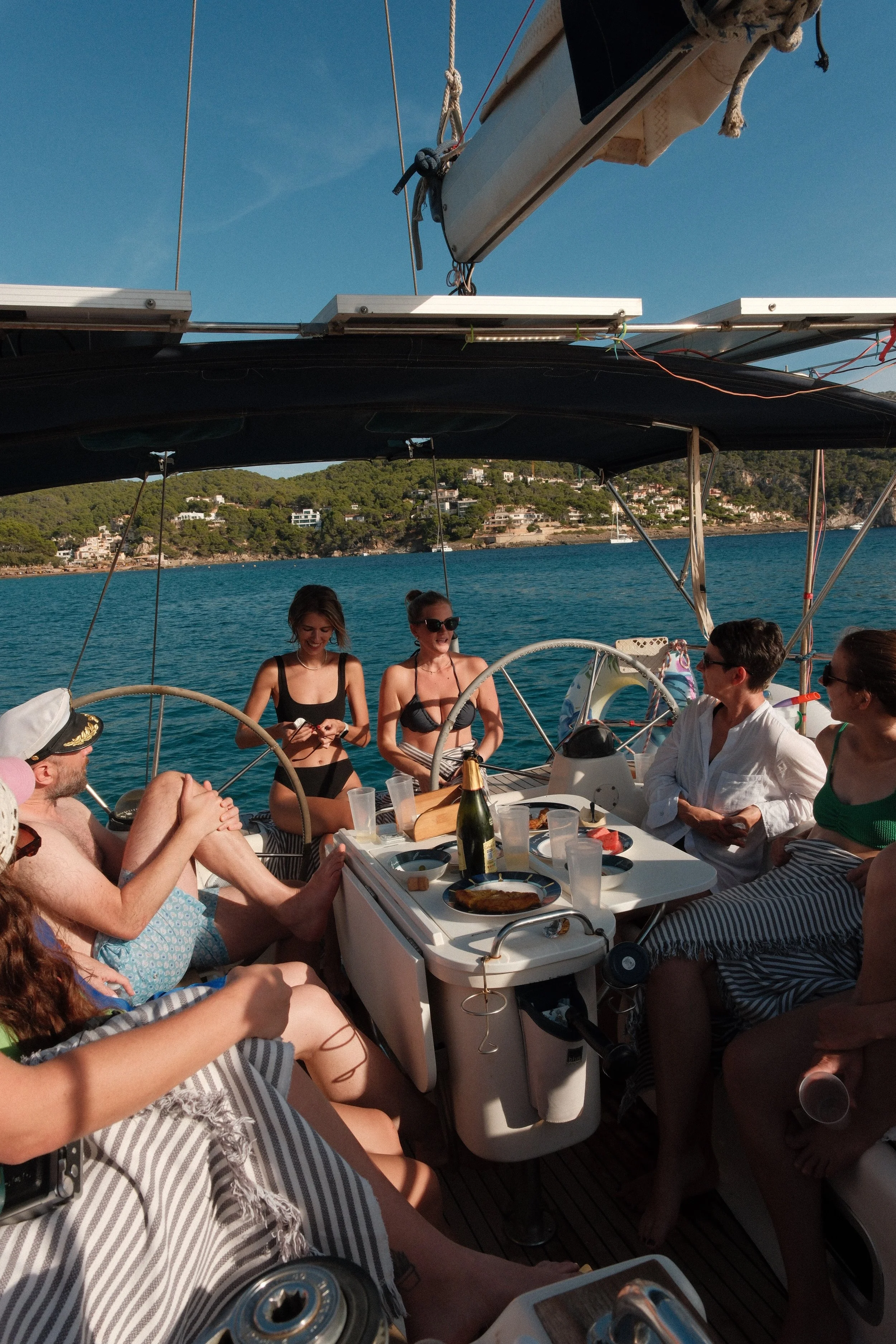 Group of people having a meal on a boat, with a scenic water and hillside background, during daytime.