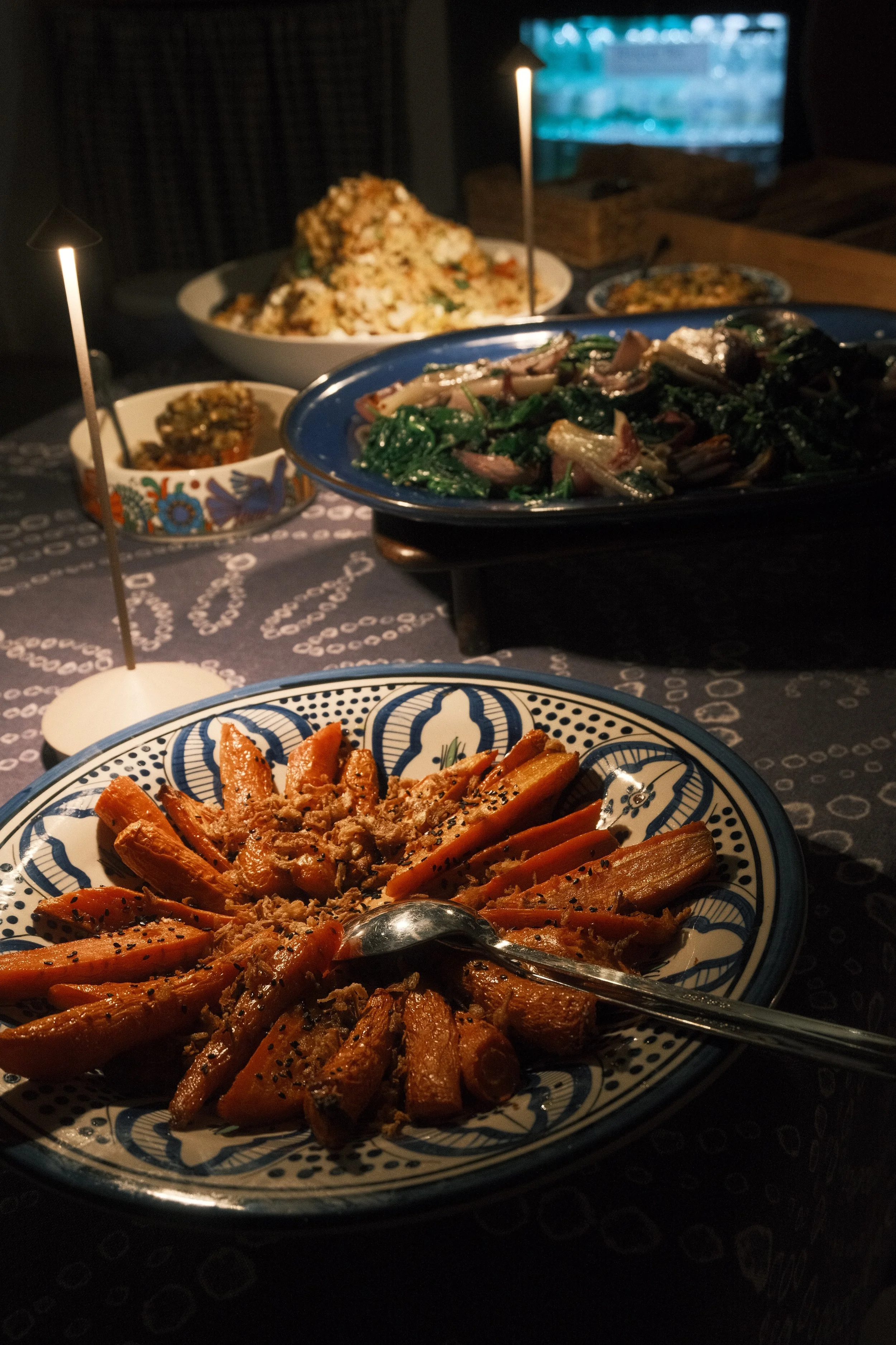 A table with various dishes including roasted carrots with herbs in a patterned bowl, a vegetable stir-fry with greens and onions on a blue platter, a bowl of rice, and smaller side dishes, set in a dimly lit room with a television in the background.