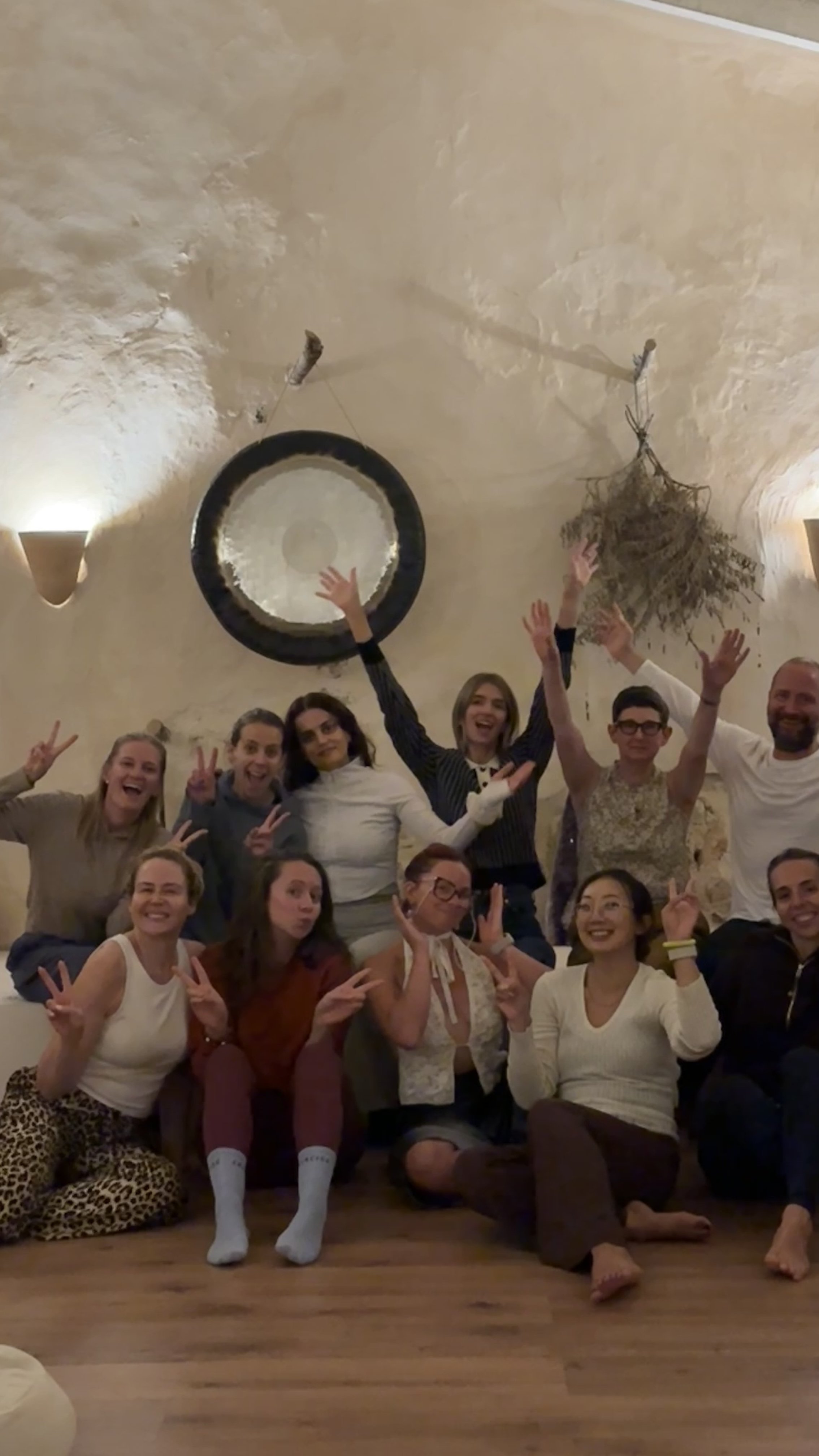 A group of people posing indoors with their hands raised, making peace signs, smiling, sitting or standing on a wooden floor under a textured ceiling.