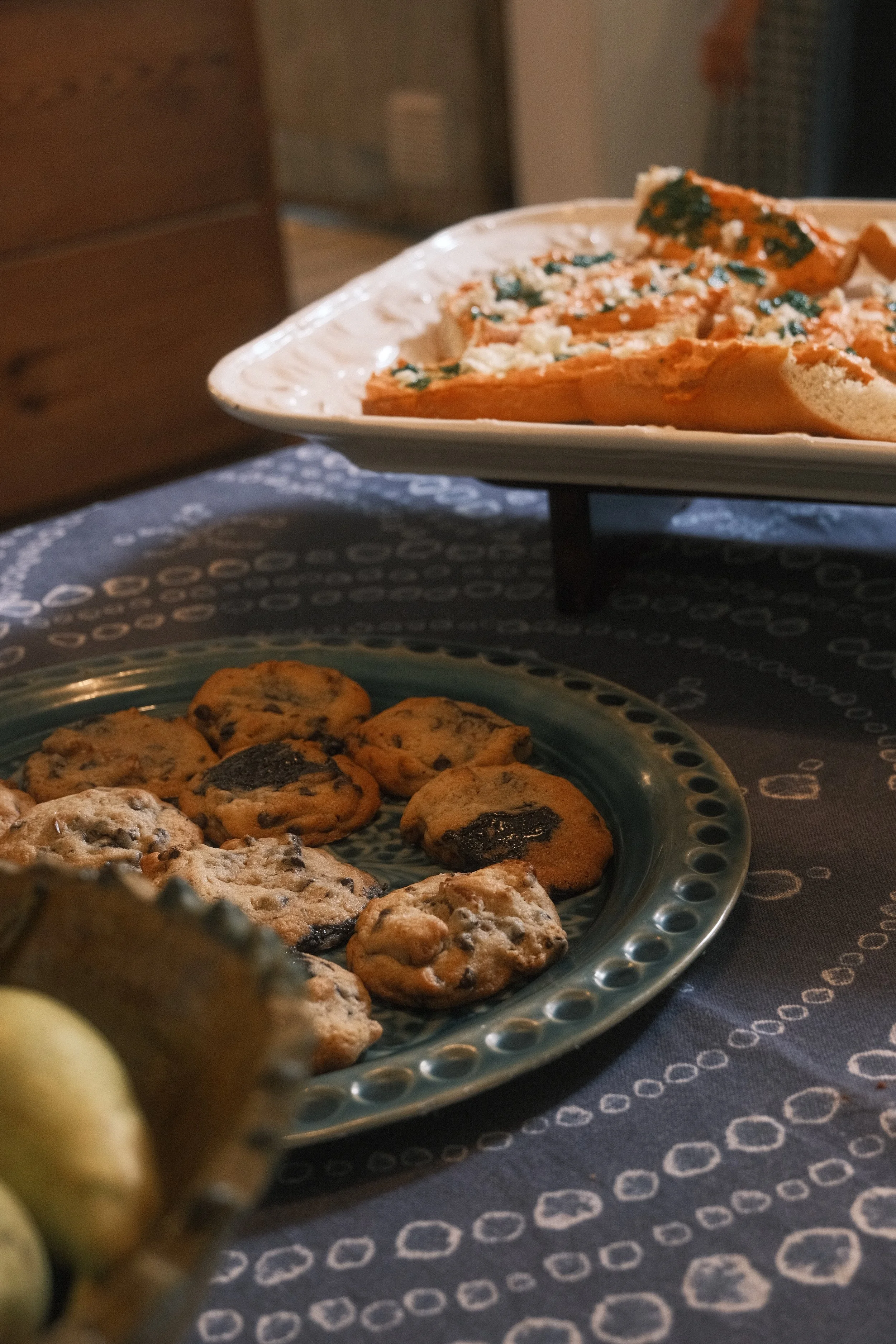 A plate of chocolate chip cookies in the foreground, with a larger serving plate of spinach and cheese stuffed bread slices in the background on a table.