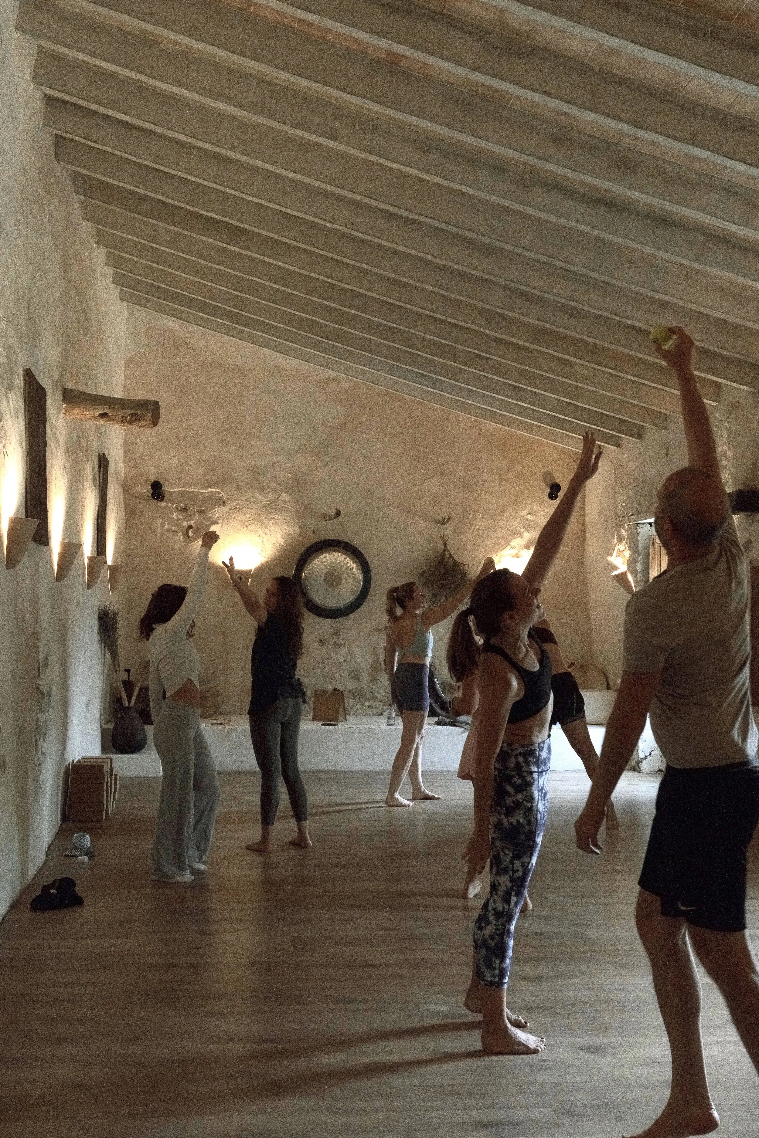A group of people participating in a yoga or meditation class in a rustic studio with exposed beams and warm lighting. They are standing on a wooden floor, some with arms raised or stretched, and are dressed in comfortable workout clothes.