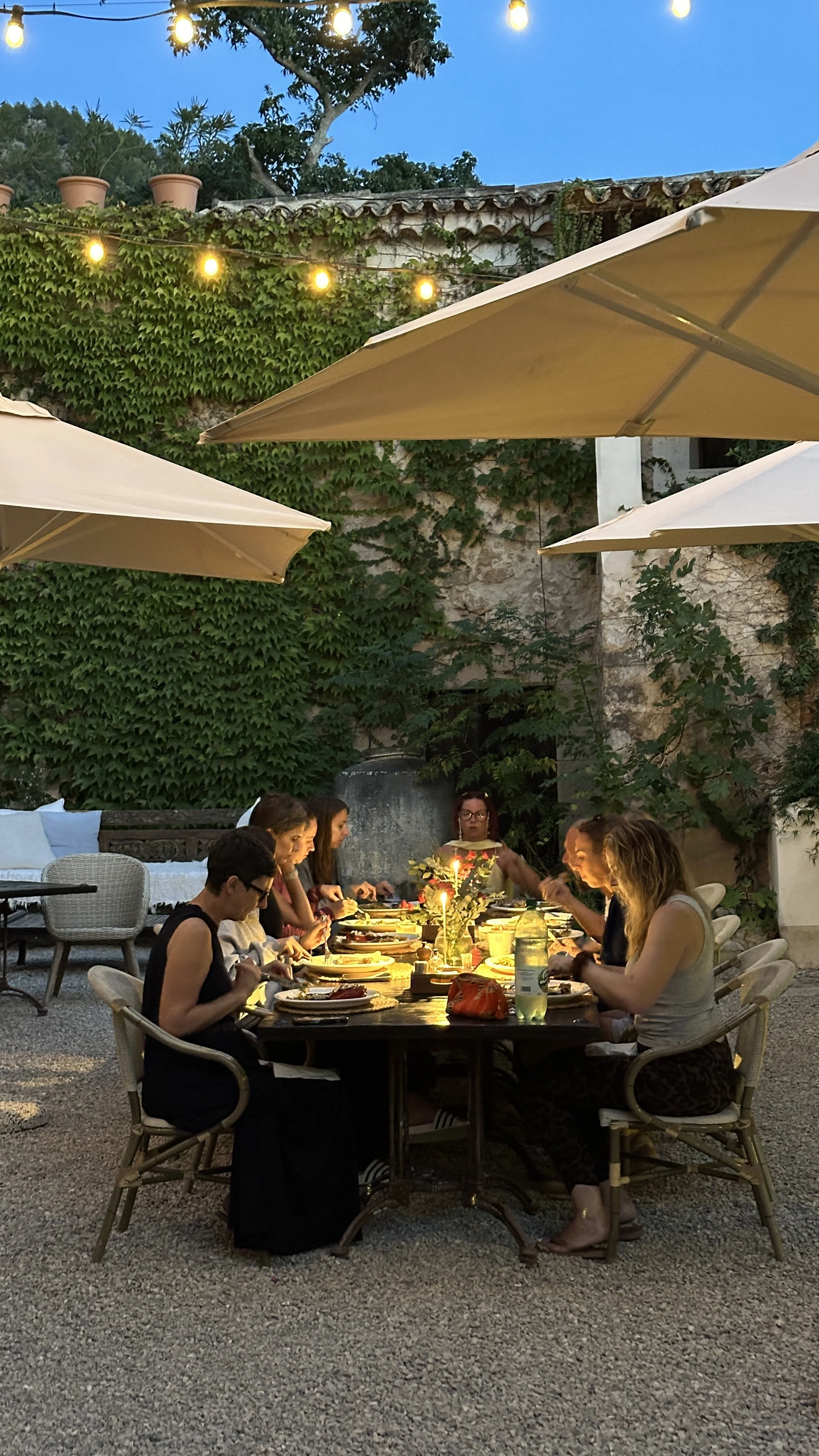 People gathered around a dinner table outside, dining under string lights and umbrellas during early evening, with a stone wall and lush greenery in the background.