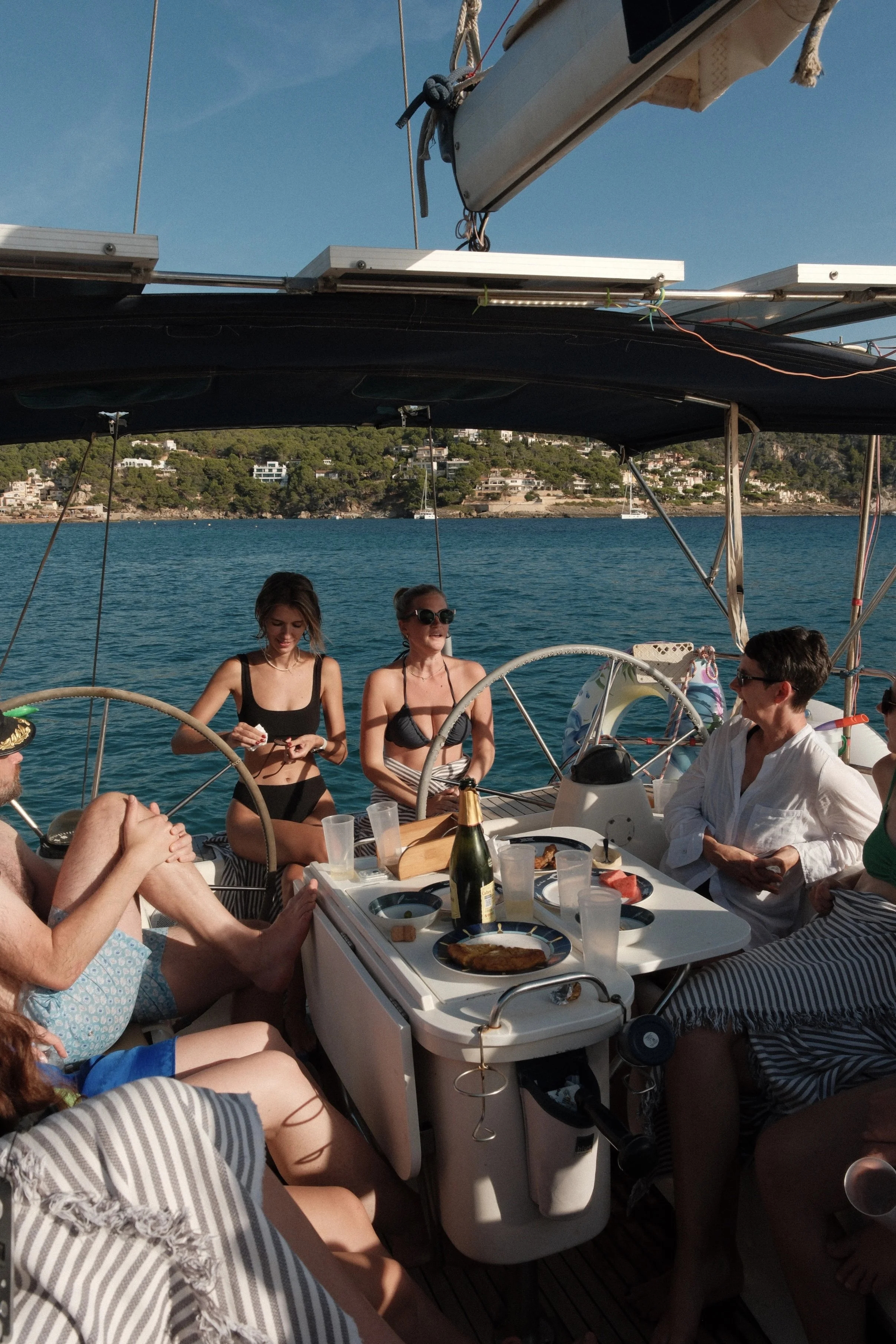 A group of people having a meal on a boat with water and coastal houses in the background.