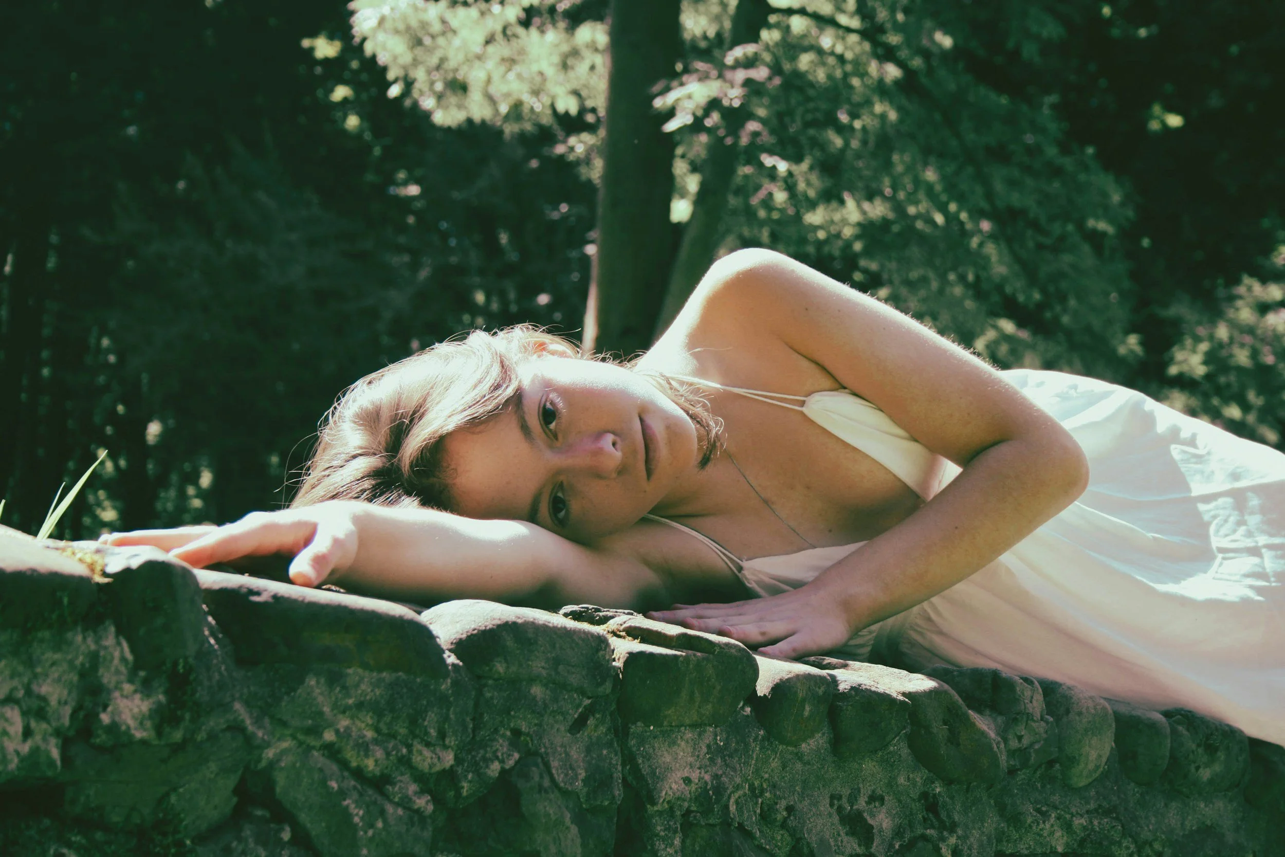 Mujer descansando en un muro de piedras en un parque, vestida con un vestido blanco y con cabello rubio, rodeada de vegetación y árboles.