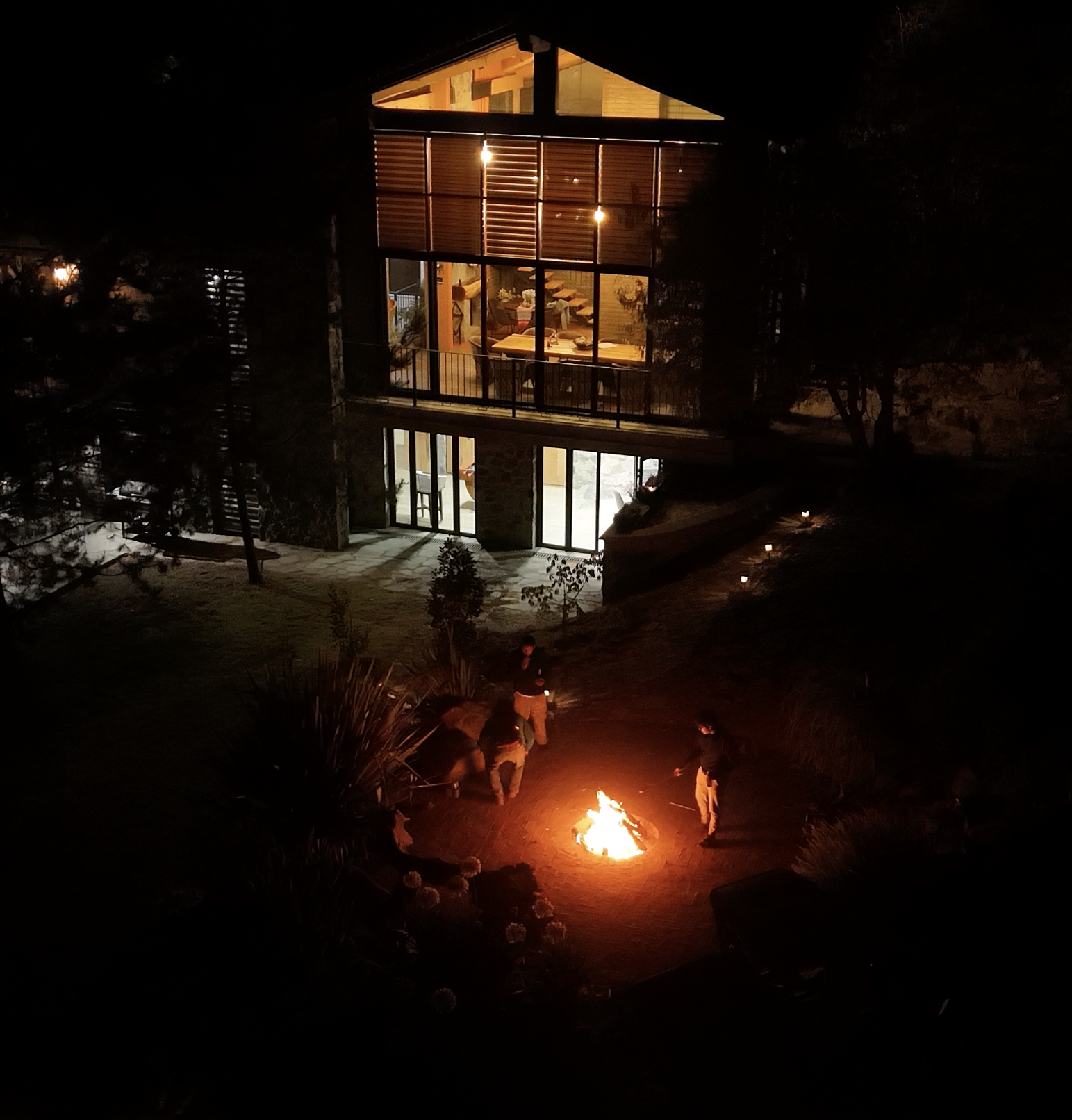 Grupo de personas alrededor de una fogata en el exterior de una casa moderna con ventanas grandes y luces cálidas, en la noche.