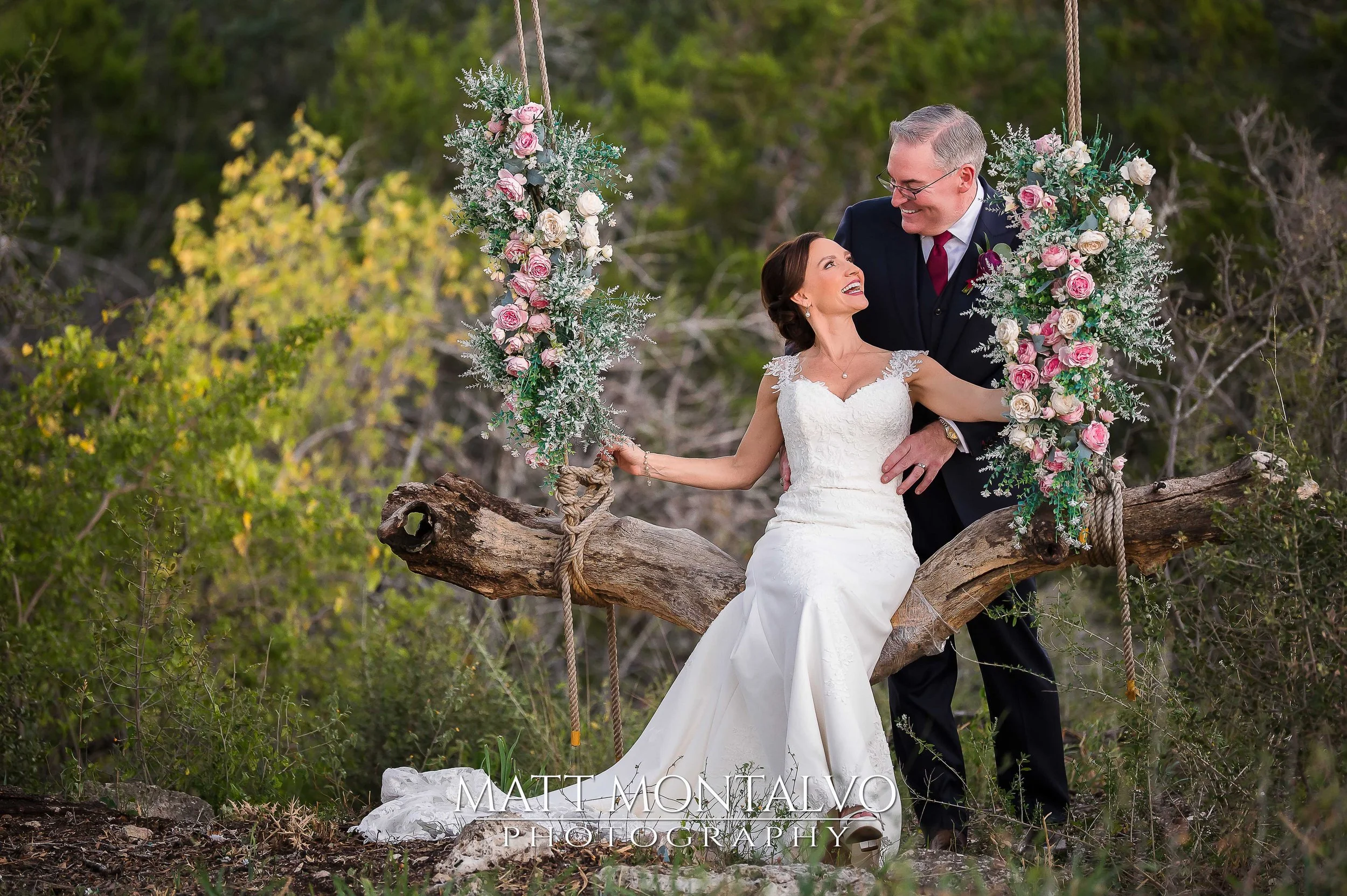 A bride and groom sitting on a large horizontal tree branch during their wedding photoshoot, surrounded by green trees and flowers, with the bride in a white wedding gown holding onto a floral swing and the groom in a dark suit looking at the bride.