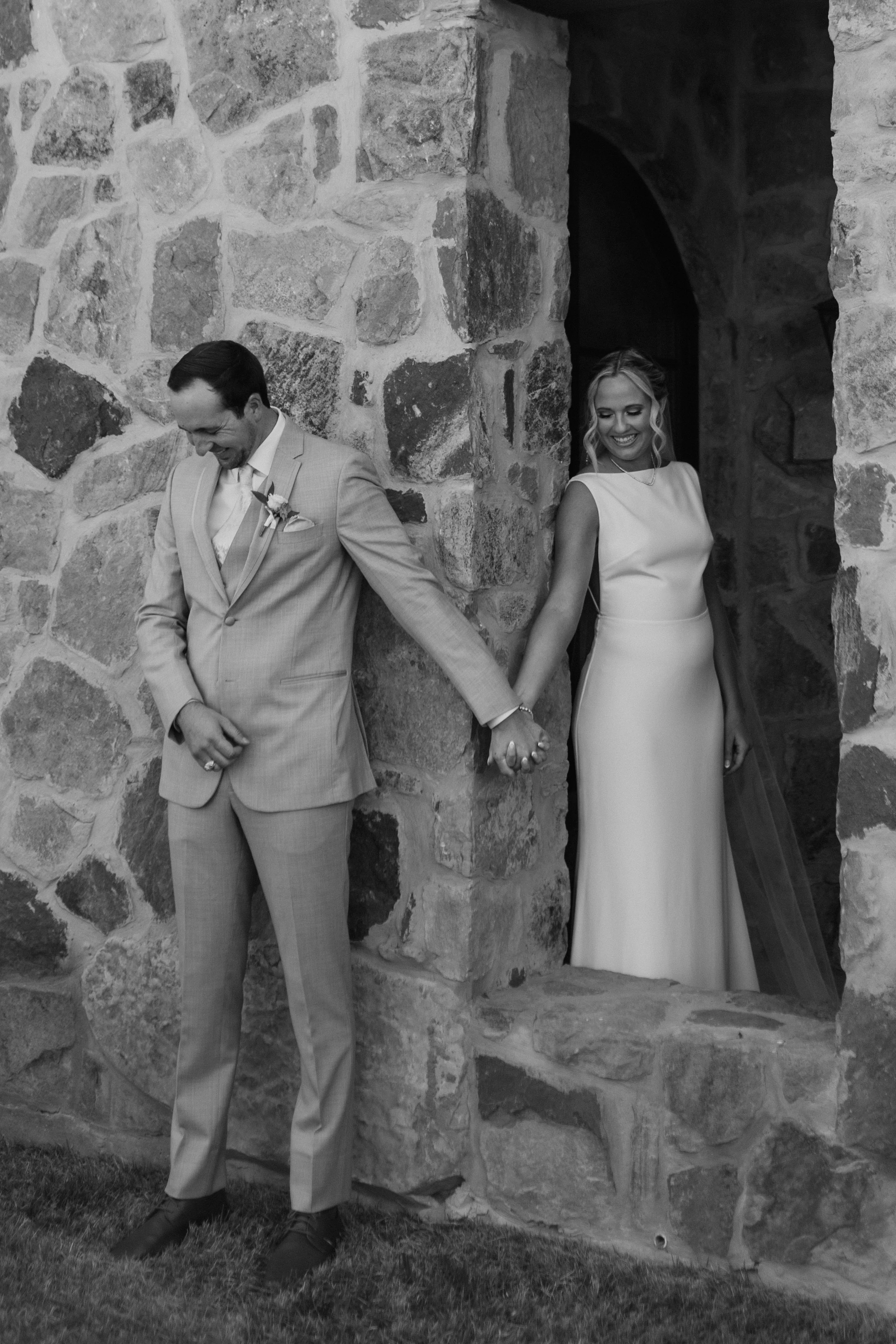 A black and white photo of a bride and groom holding hands, standing near a stone wall. The groom looks down and is dressed in a light-colored suit with a boutonniere, and the bride, smiling, is dressed in a sleeveless gown, with her hair styled in soft curls.