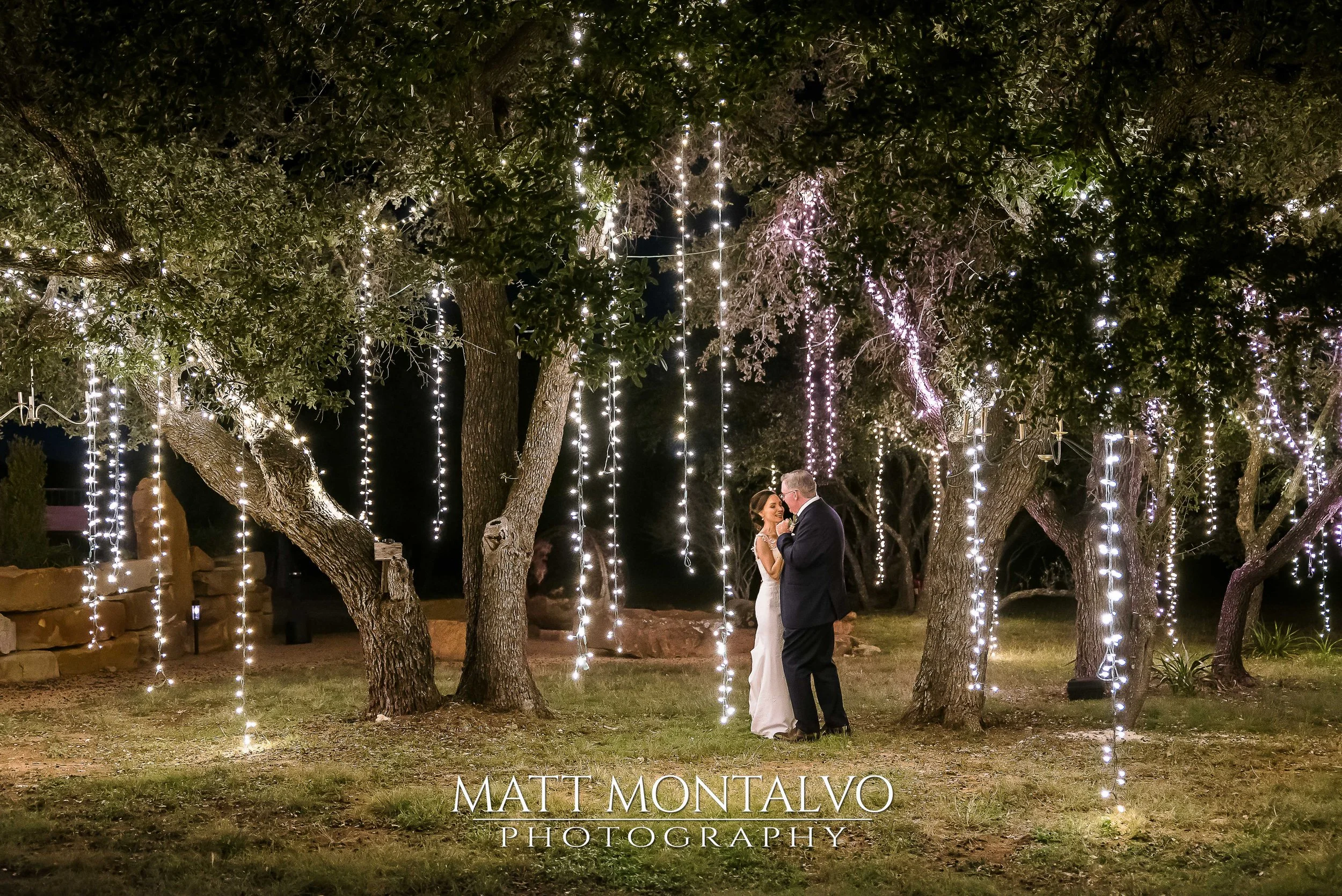 A bride and groom dancing under a large tree decorated with hanging lights at night. The scene is illuminated by string lights creating a romantic atmosphere. The photo is credited to Matt Montalvo Photography.